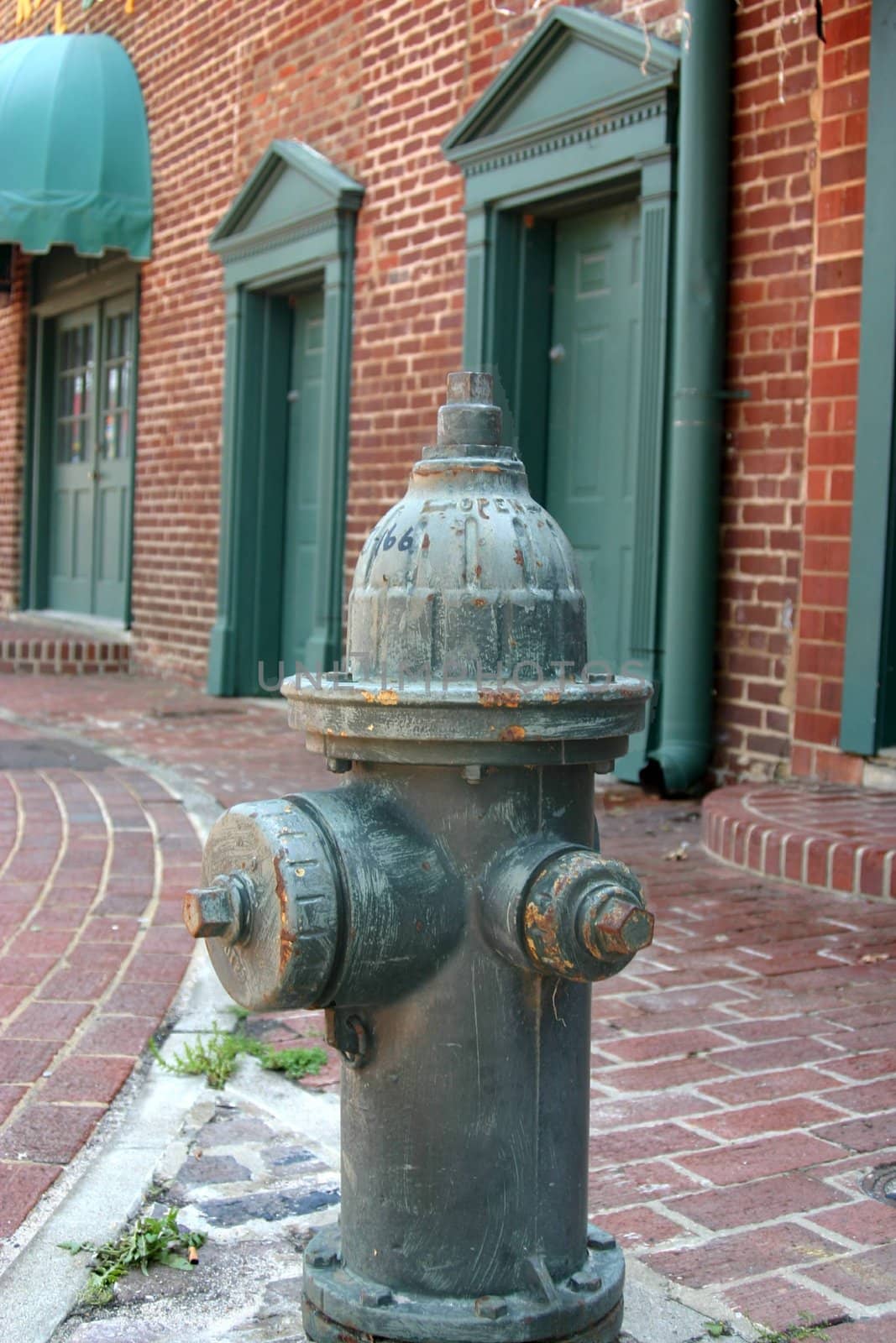 This is a green fire hydrant in an old commercial district on a brick paved street. 