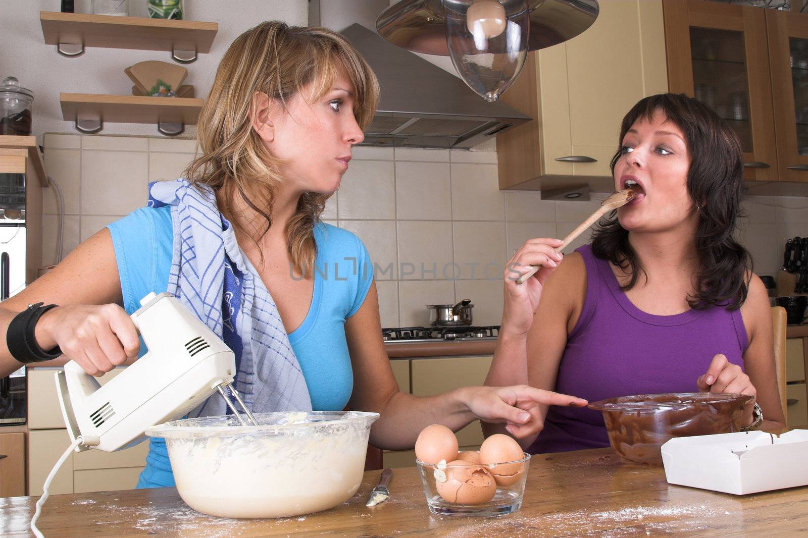 Pretty girls cooking together in the kitchen making cake and brownies. One girl is licking the chocolate of the spoon