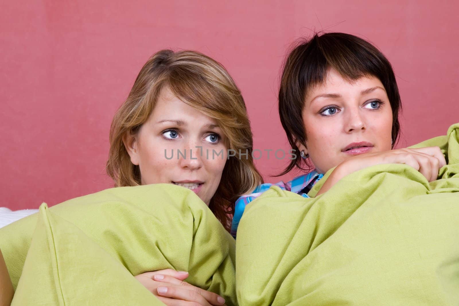 Two friends watching a scary movie on the couch hiding behind big pillows