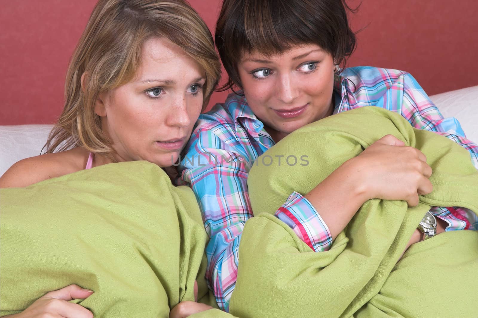 Two girls watching a scary movie together while hiding behind big pillows