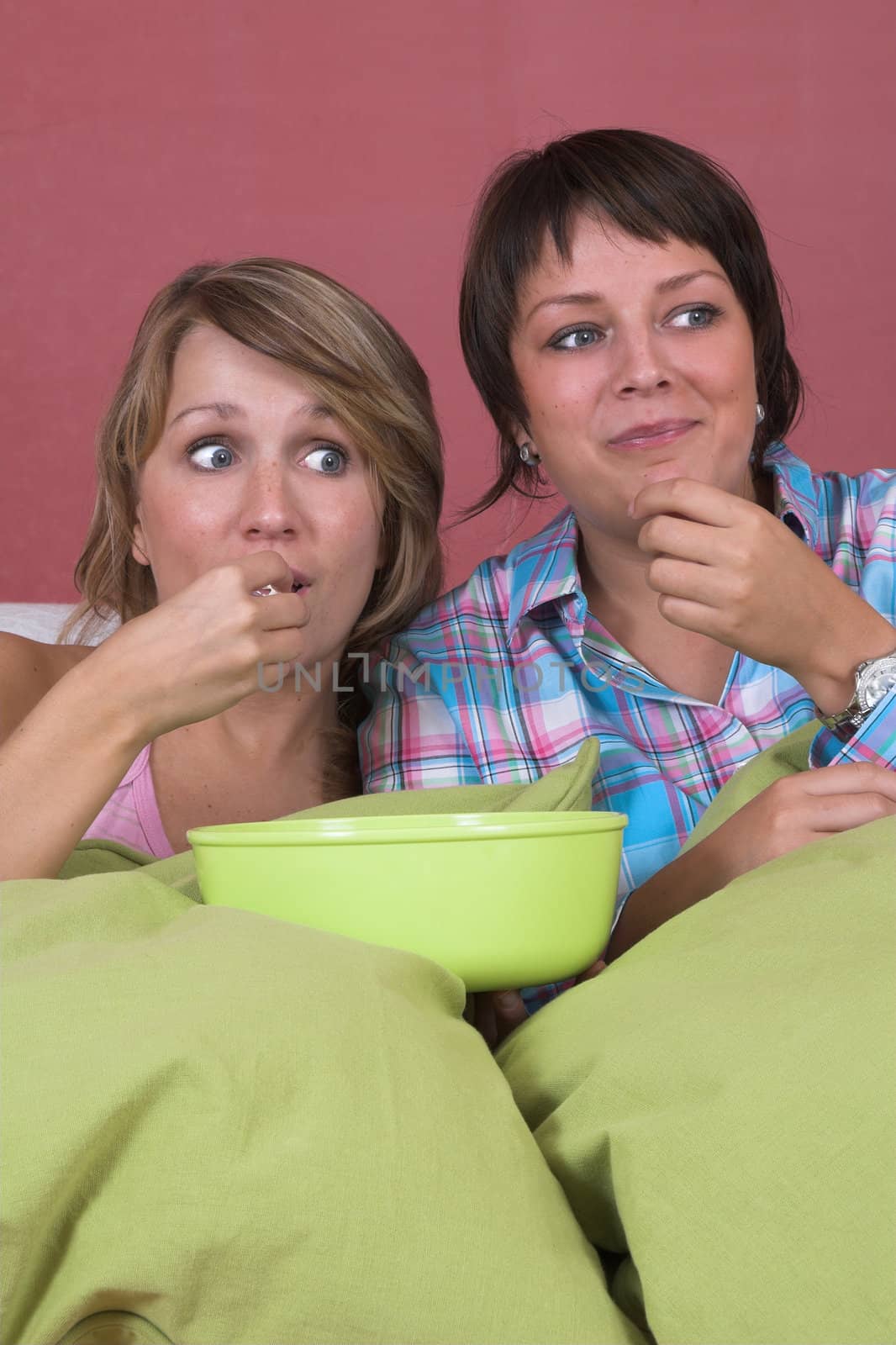 Two girls on the couch together watching a movie while eating popcorn and looking surprised