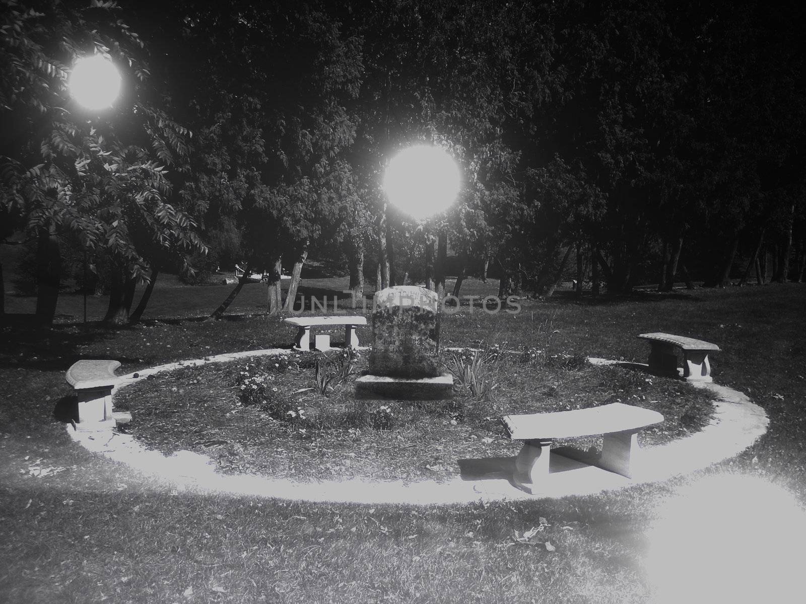 black and white photo of a gravestone surrounded by a ring and benches. Around the gravestone float three balls of ghostly light.