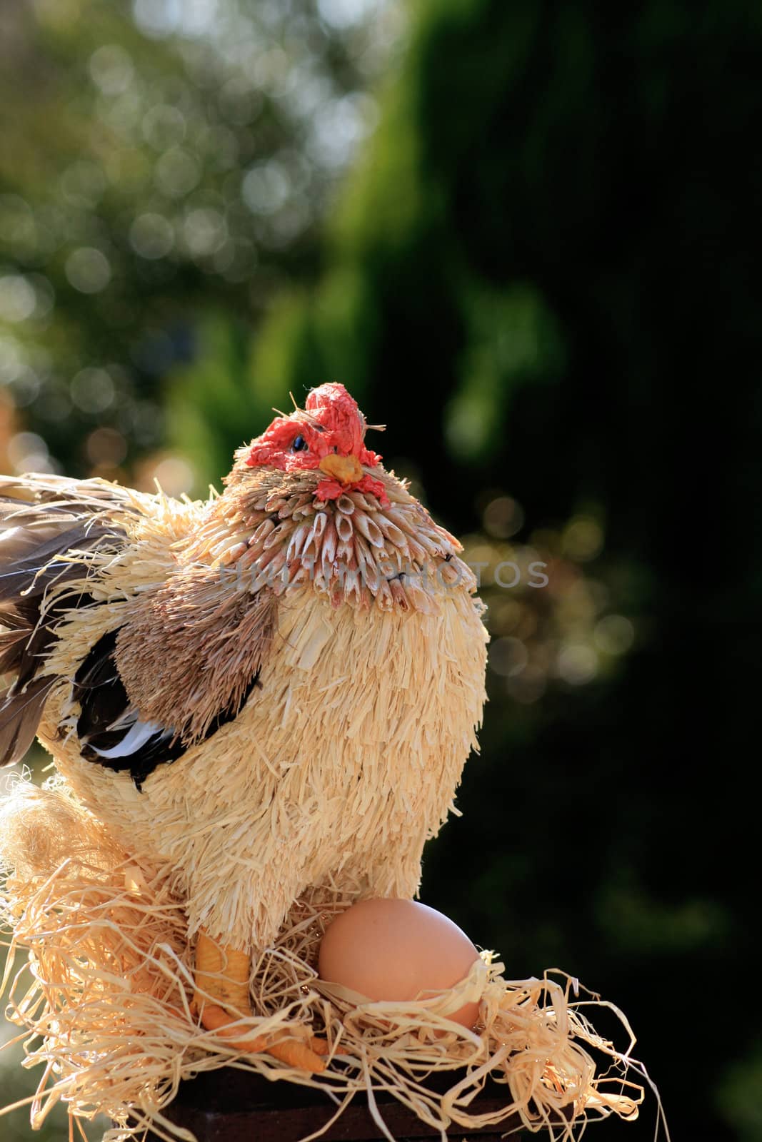 A hand made straw and string constructed chicken/rooster perched atop a wooden post with an egg.