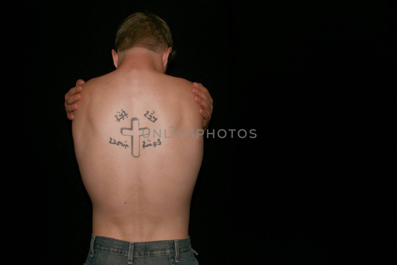 A topless man hugging himself, his back showing a crucifix tattoo.
