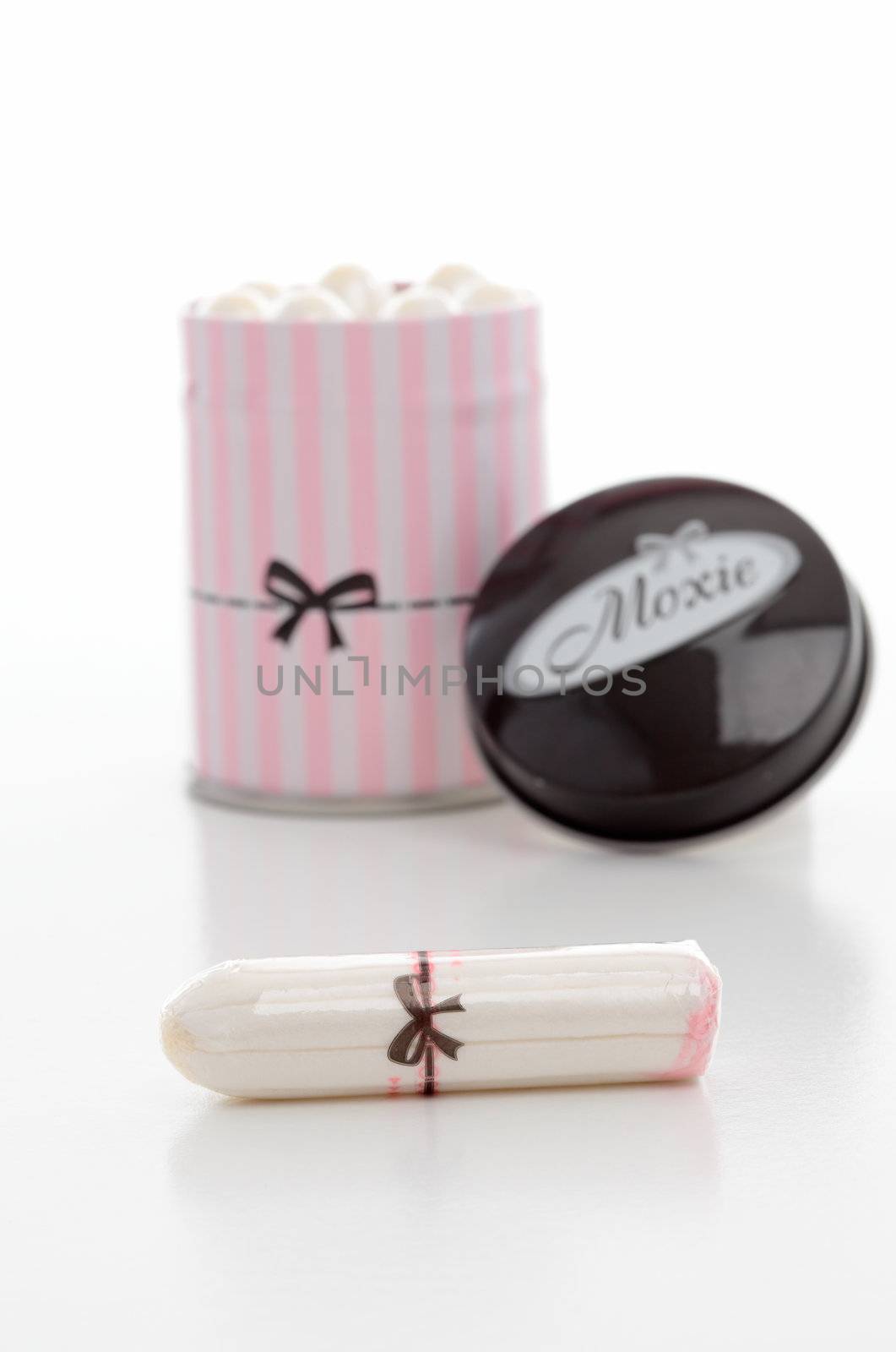 A tin of Moxie tampons and one single tampon in the foreground.  Focus to foreground.  White backdrop.