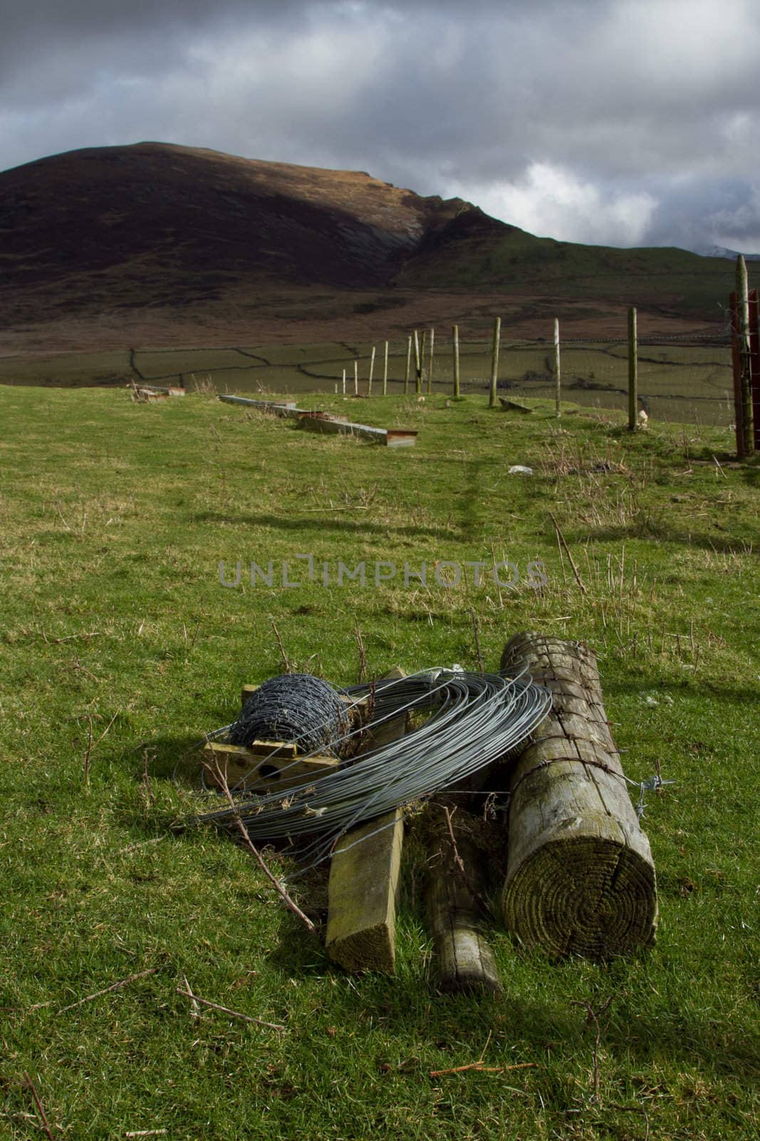 Agricultural landscape with wooden fence posts a coil of wire and a spool of barbed wire on green grass with a hill in the background.