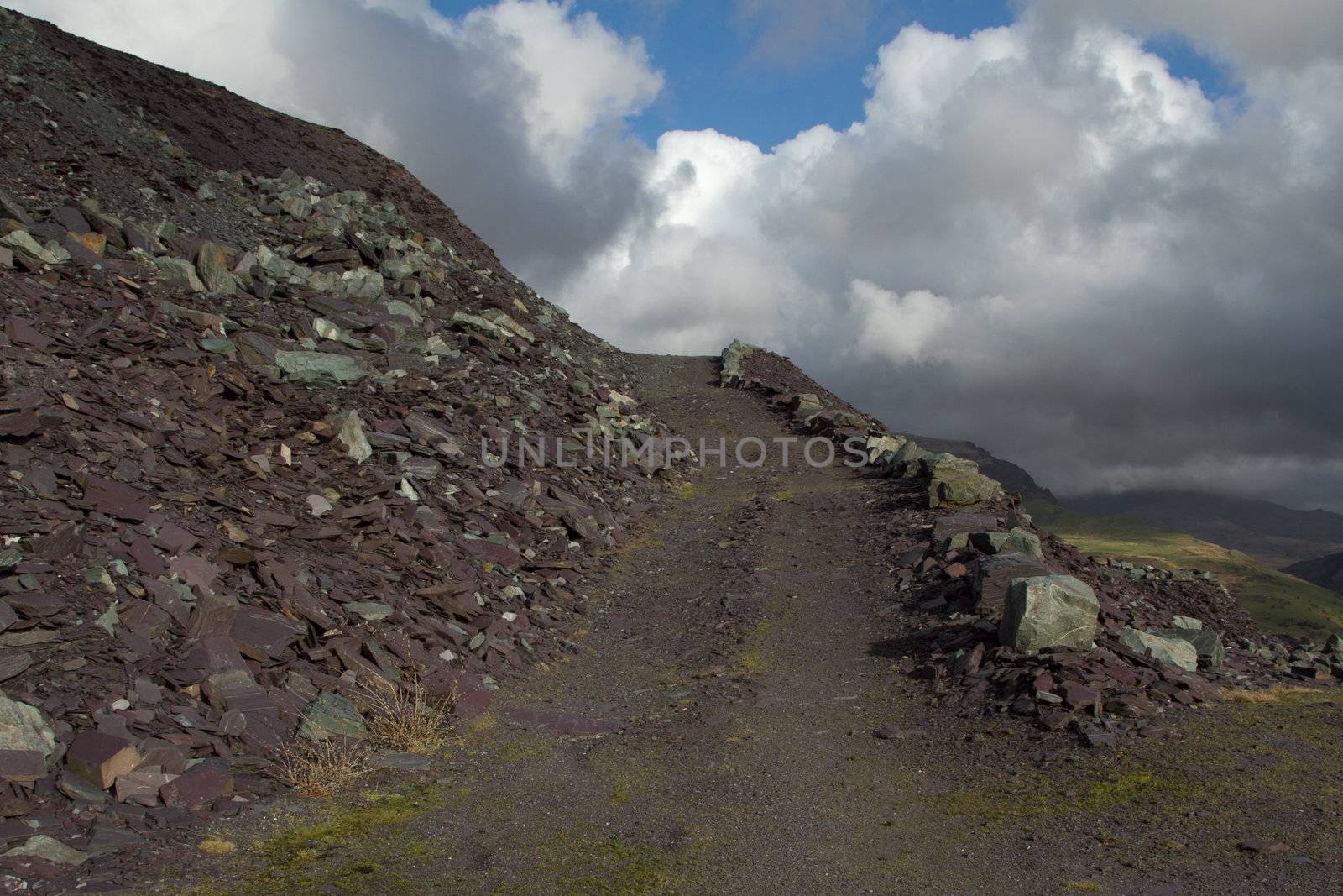 Quarry land management, with slate waste in a heap and a track leading into the distance, lined with boulders leading to a clean blue cloudy sky.