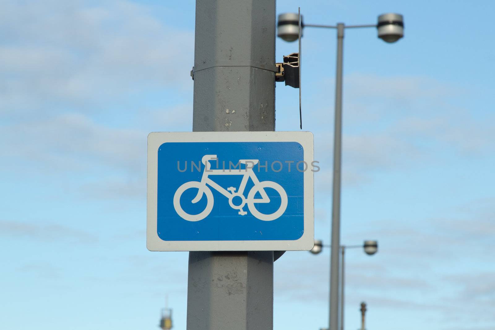 A sign, blue with a white border and a symbol of a bicycle, attached to a lamp-post with a blue sky in the background.