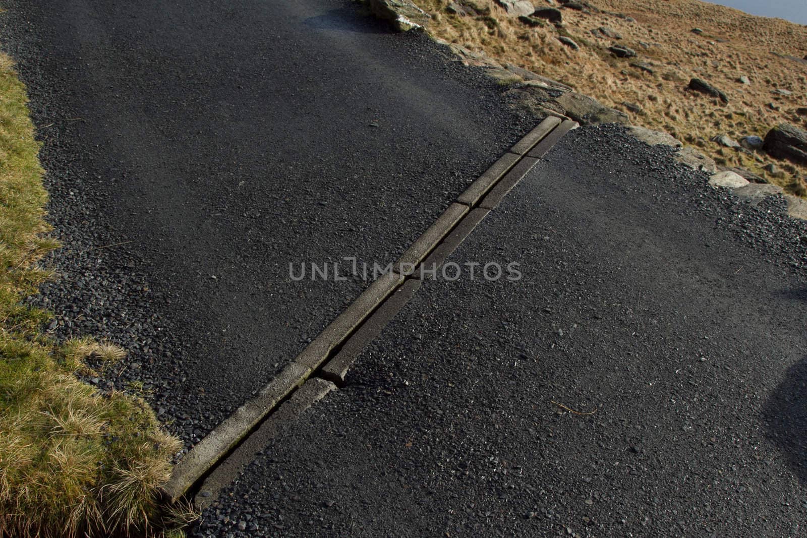 A tarmac footpath with stone curbs and grass with a sectioned drainage system spanning the width.