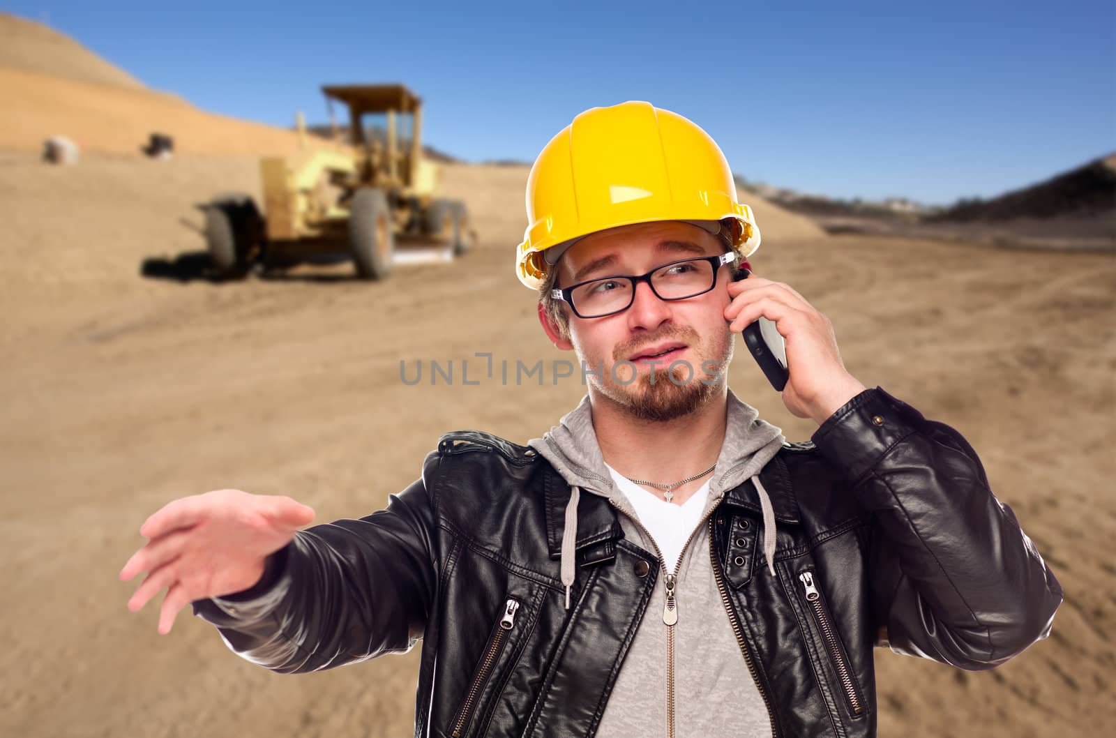Young Cunstruction Worker on Cell Phone in Dirt Field with Tractor in the Background.