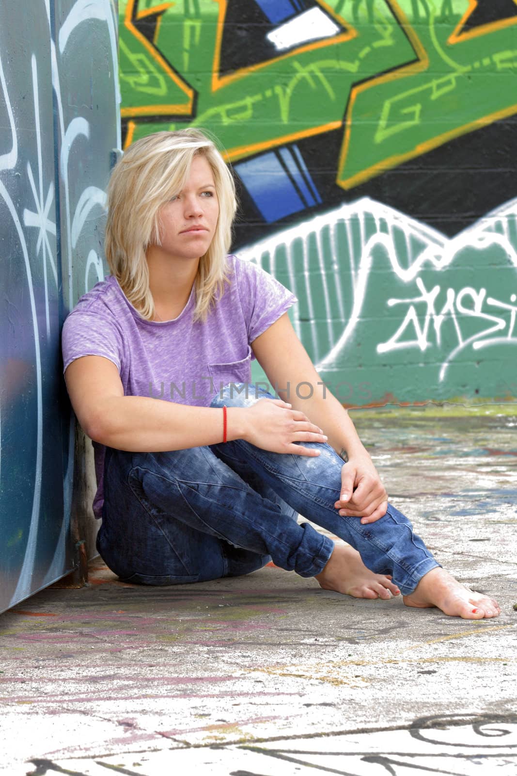 A lovely young blonde leans against a graffiti-covered garbage dumpster with a graffiti-covered wall behind her.