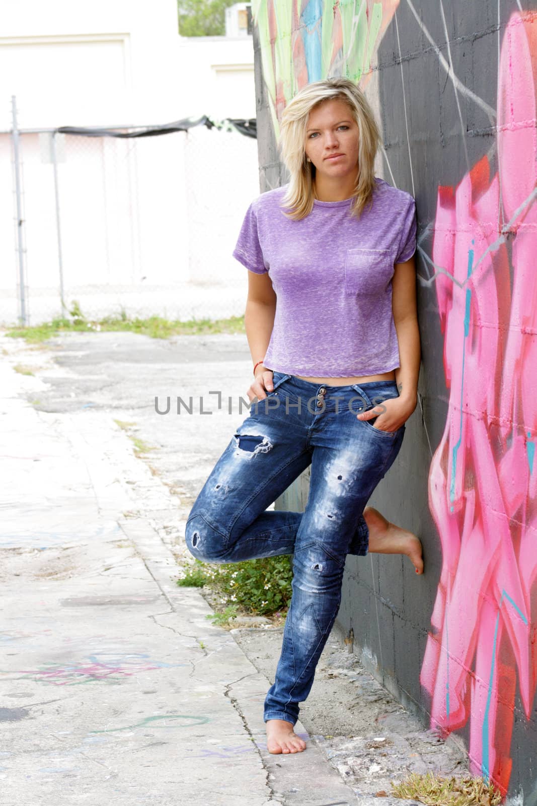 A lovely young blonde leans against a graffiti-covered wall in an urban environment.