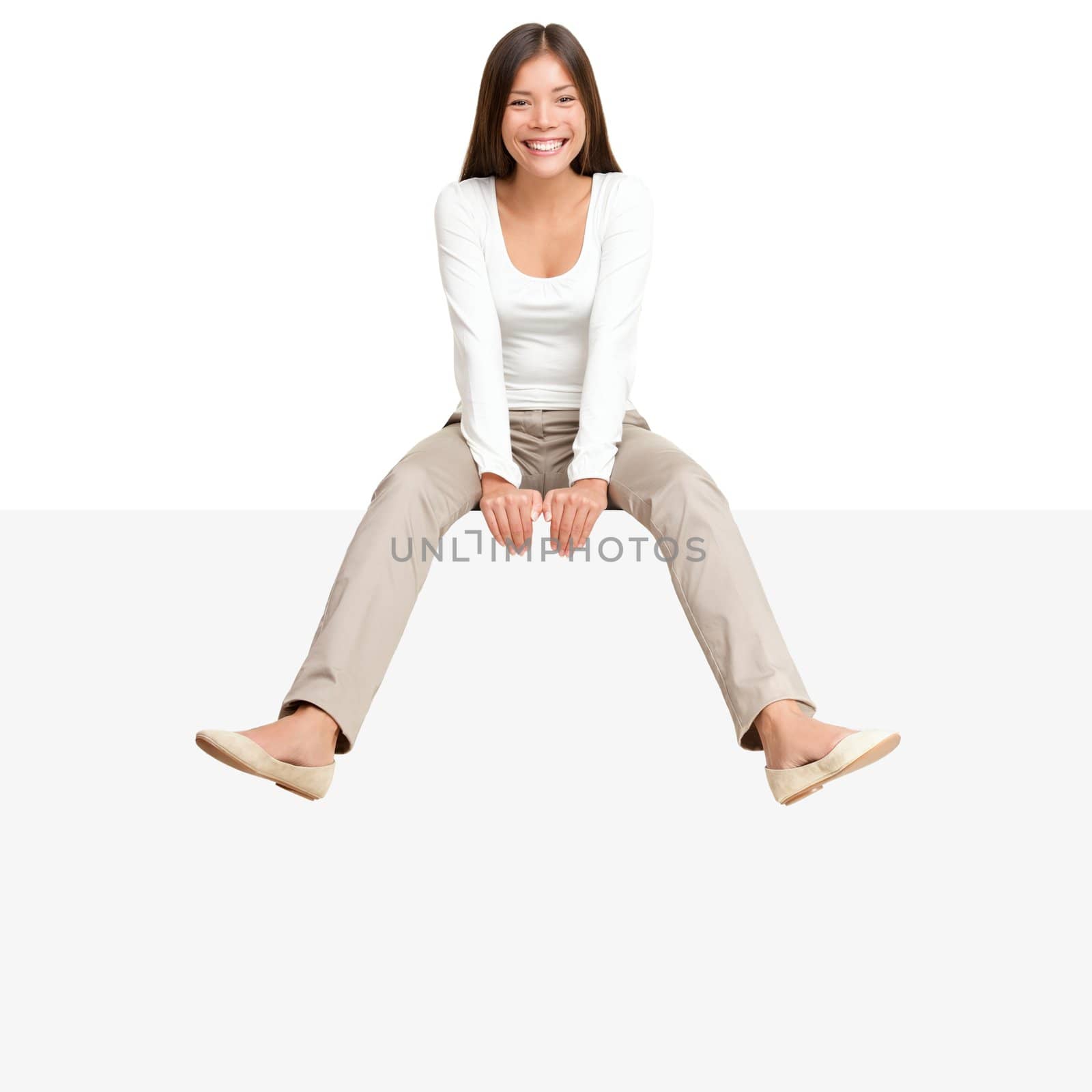 Pretty girl sitting casual on big blank billboard poster sign with lot of copy space. Smiling asian caucasian young woman model. Isolated on white background