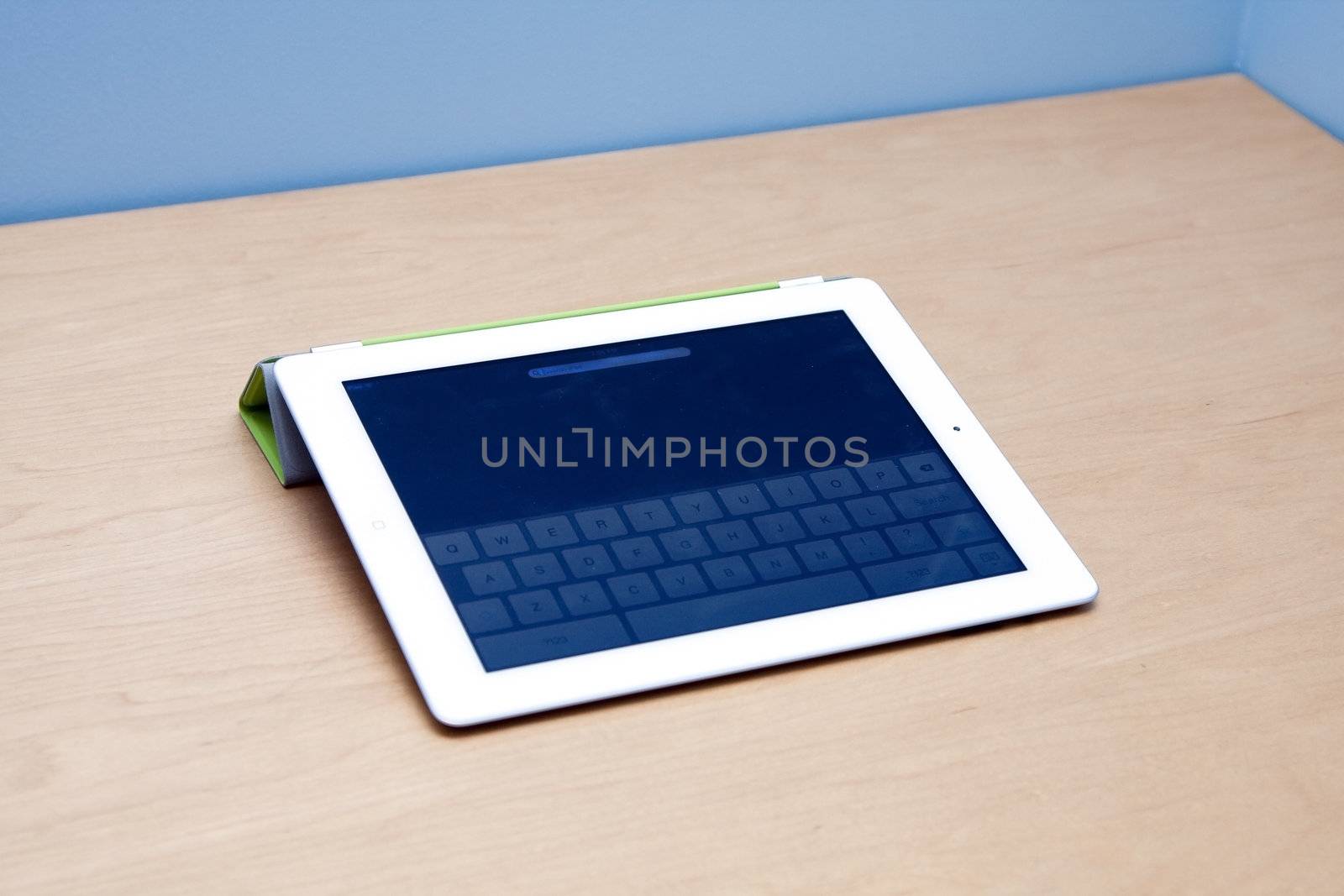 The new iPad 2 from Mac Apple, on a wooden desk and blue background with visible keypad touchscreen and forward facing camera.