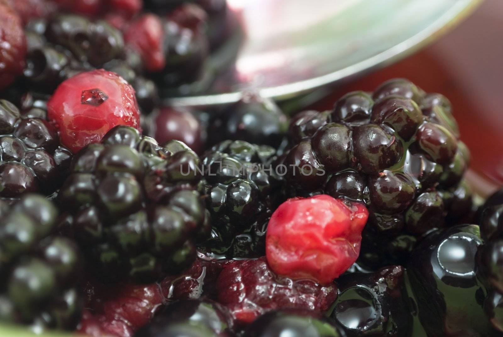 Closeup shot of summer fruits in a bowl, comprising blackberries, raspberries, redcurrants and blackcurrants in their own juice.  A spoon scoops some berries in soft focus in the background.