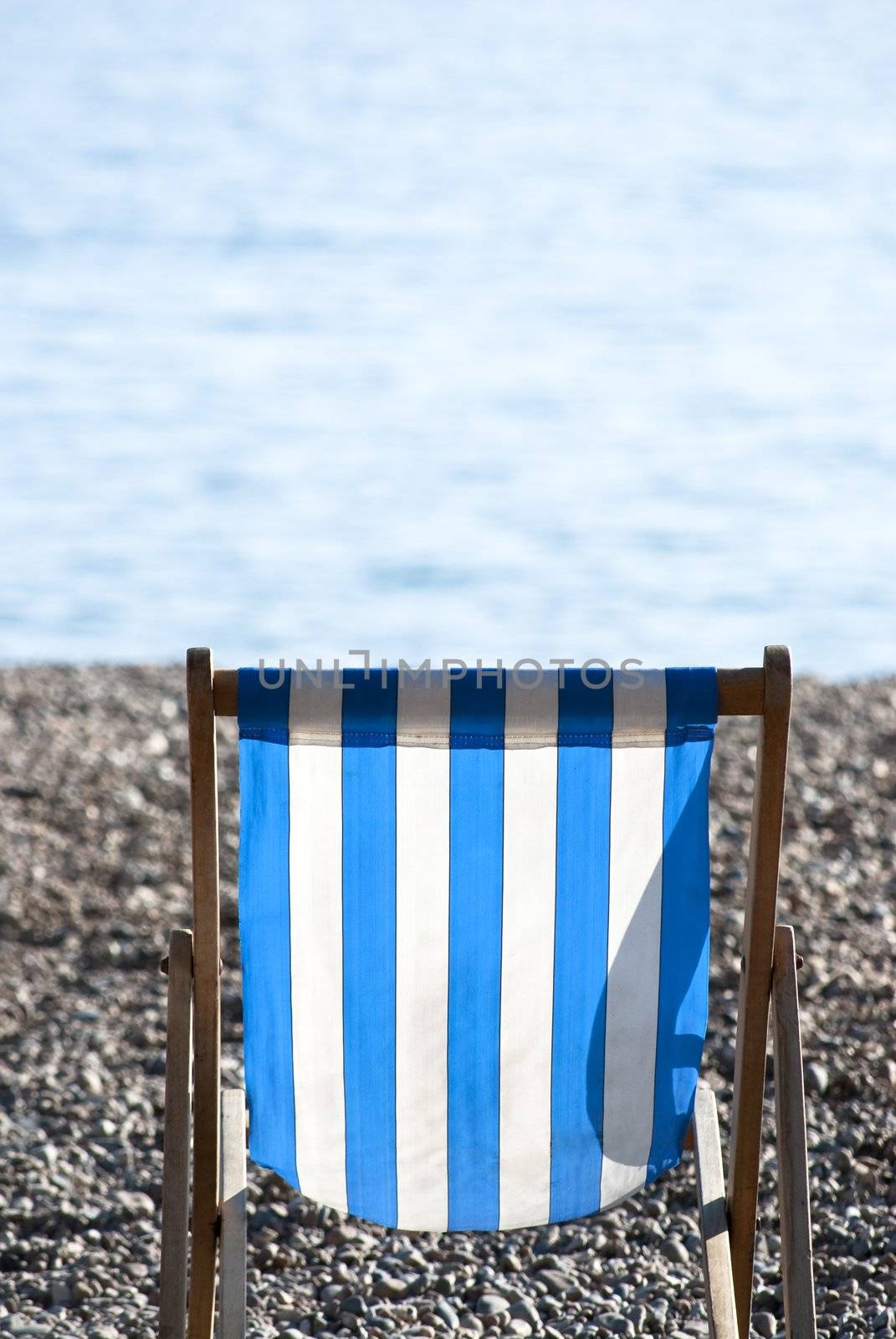 A solitary, empty, blue and white striped deckchair sitting on a pebble beach on a sunny day, with sea in soft focus in the background.