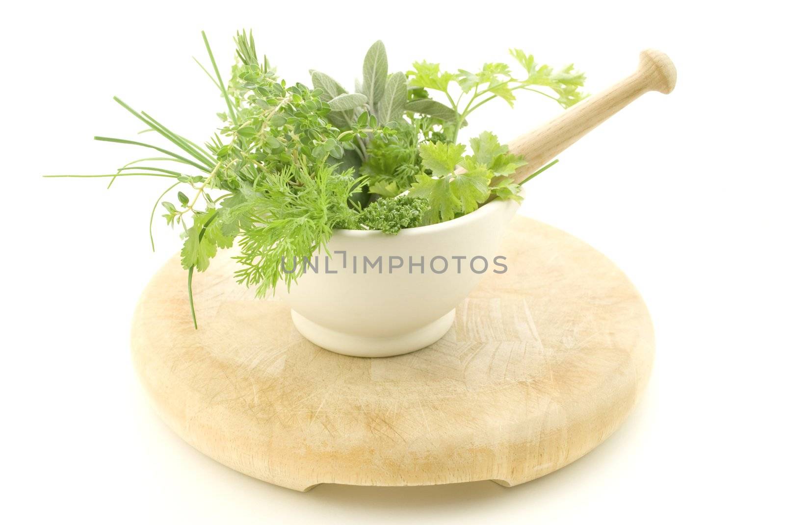 A selection of herbs inside a cream mortar with pestle, standing on light wood chopping board which shows signs of wear.  Isolated against white background.