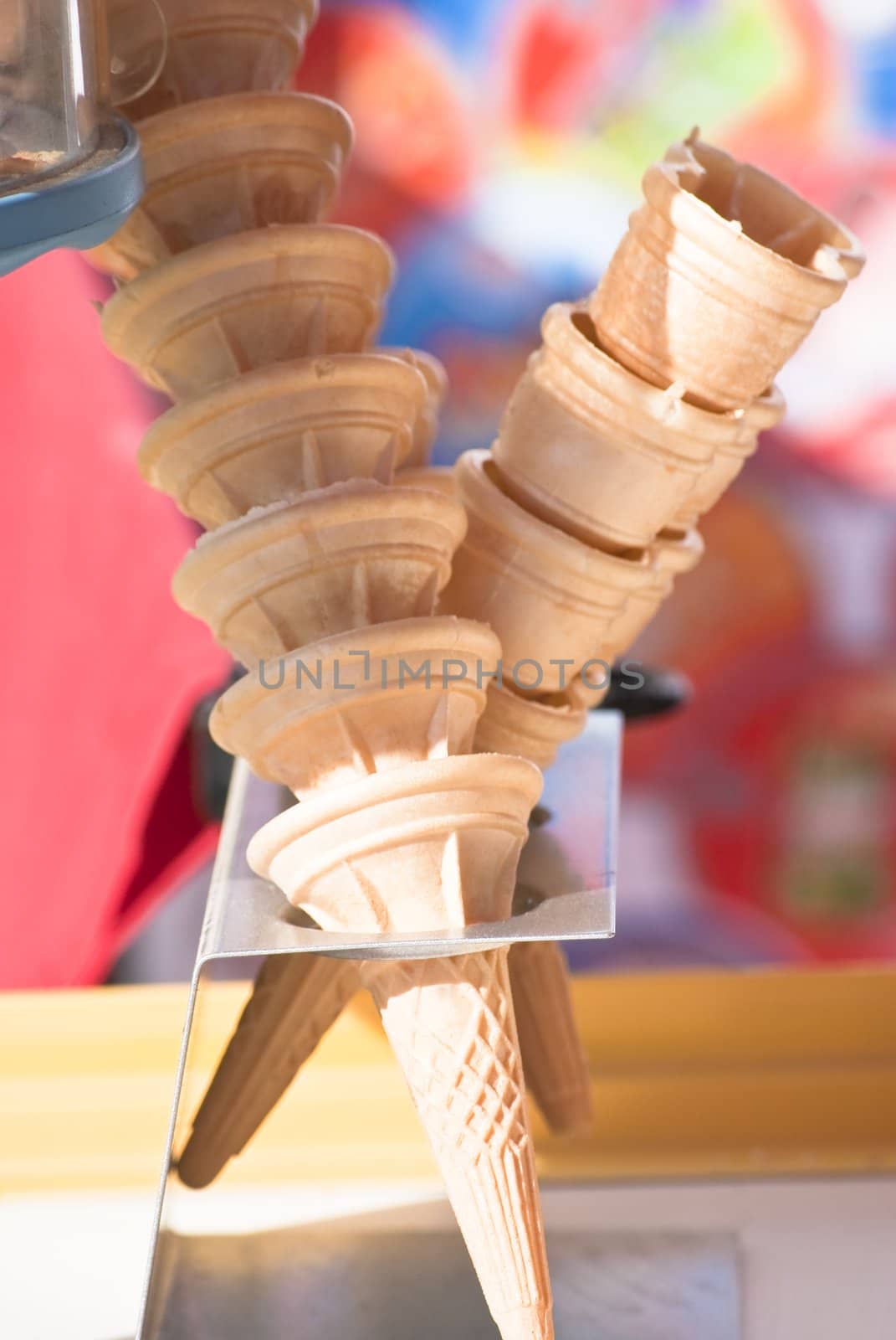 Single and double ice cream cones stacked on a seller's ice cream stand.  A mixture of strong sunlight and cool shade illustrates a hot, sunny day.  Soft focus background of bright colours: red, blue, green, white.