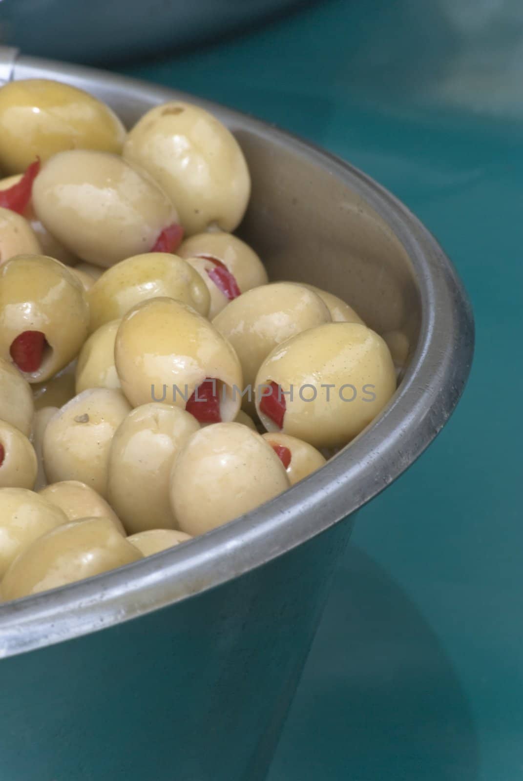 Green olives, stuffed with red chilli peppers, in a metal bowl on a teal coloured tablecloth.  Shot in daylight.  Vertical orientation (portrait).