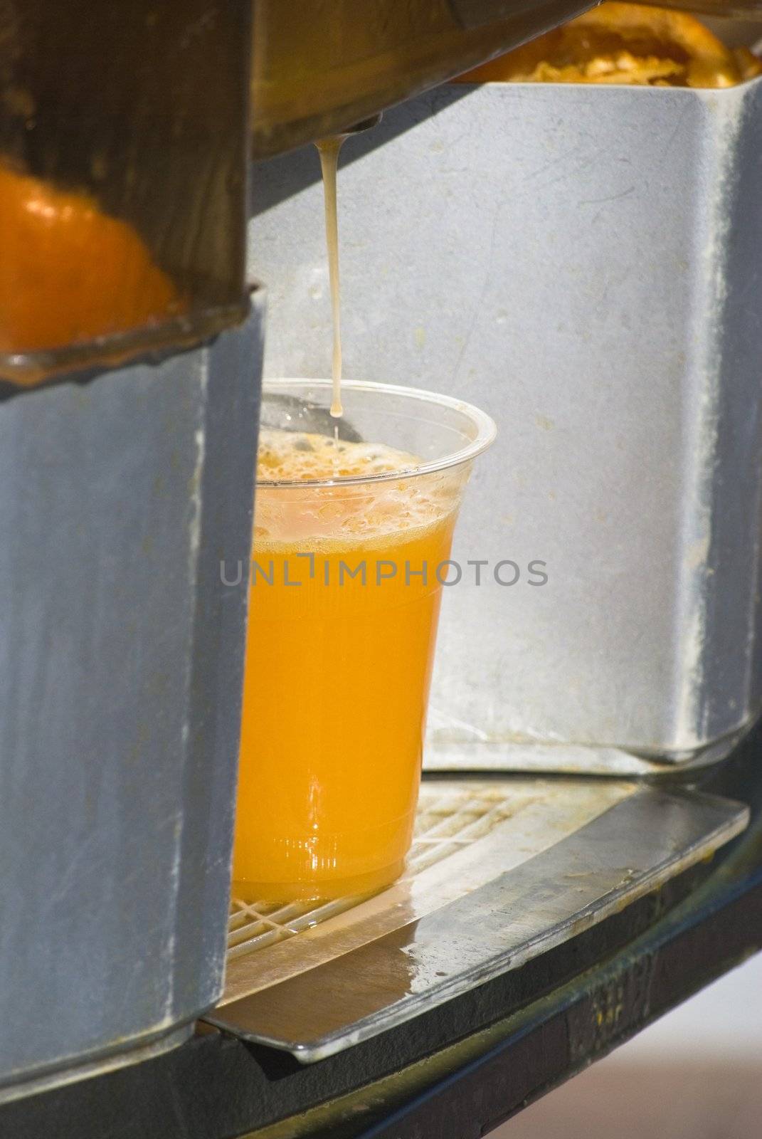 Fresh orange juice dripping into plastic shop from a juice dispenser, in strong outdoor sunlight.  Discarded peel and pulp just visible in container in the background.