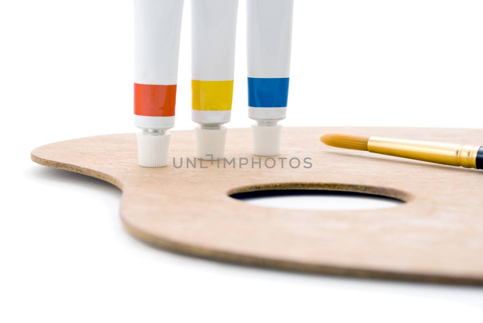 Three tubes of paint (primary colours) standing on edge of wooden palette with thumb hole and paintbrush tip visible.  Closeup shot, cropped in camera so only leftmost portion of palette is visible.  Isolated against white background.