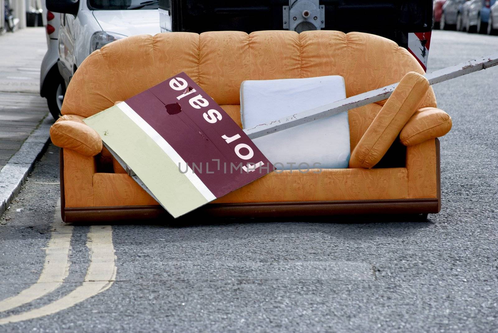 An orange sofa in a road with a For Sale sign thrown on to it.  Double yellow lines indicate that parking is not allowed.  Parked vehicles visible in the background.