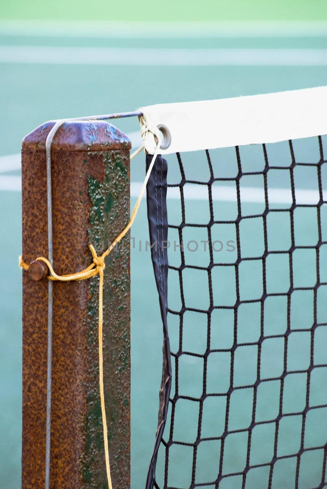 Close shot of tennis court net attached to rusting metal post with yellow and grey ropes.  Tennis court is out of focus in background.