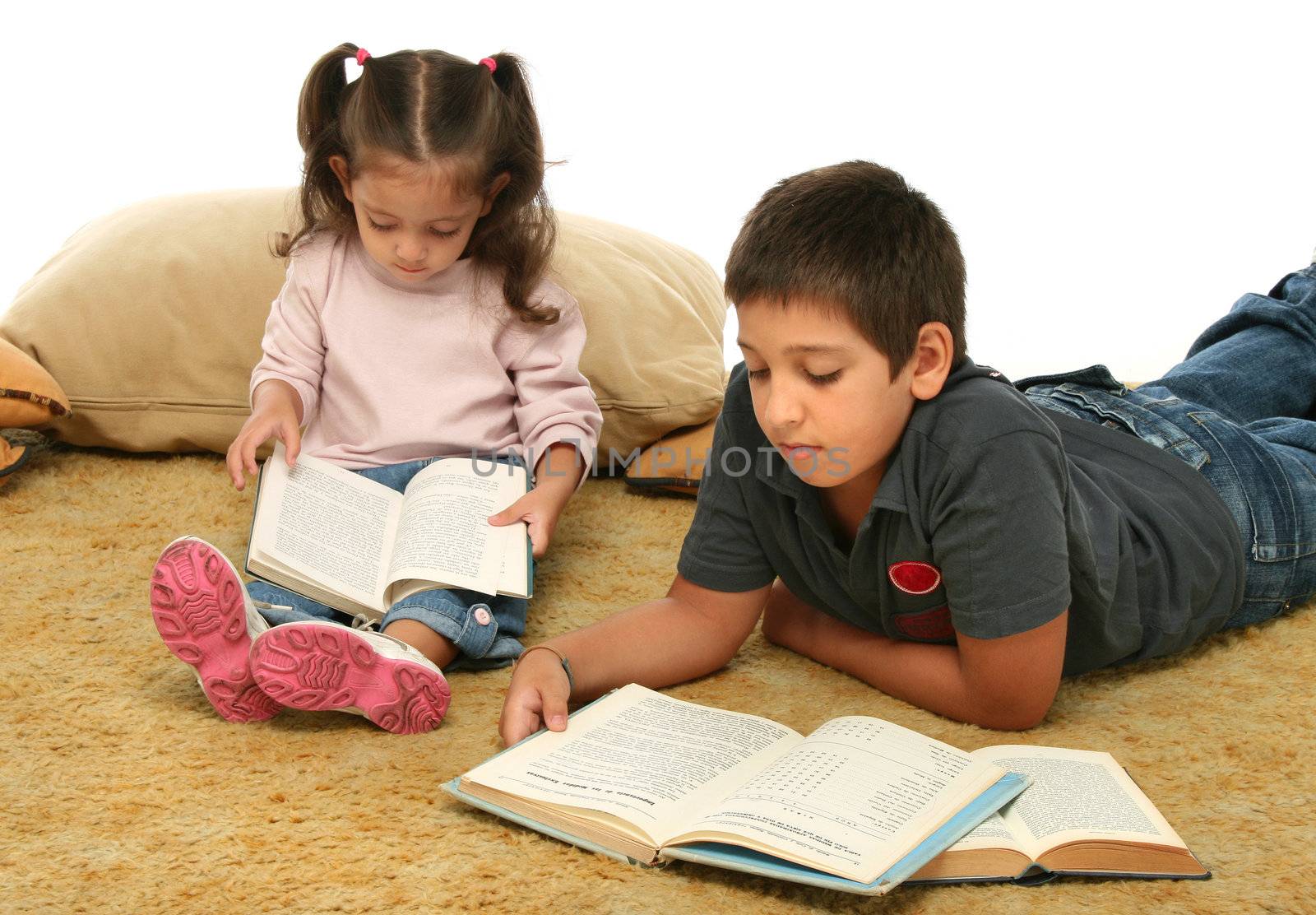 Brother and sister reading books over a carpet. They look interested and concentrated. 
