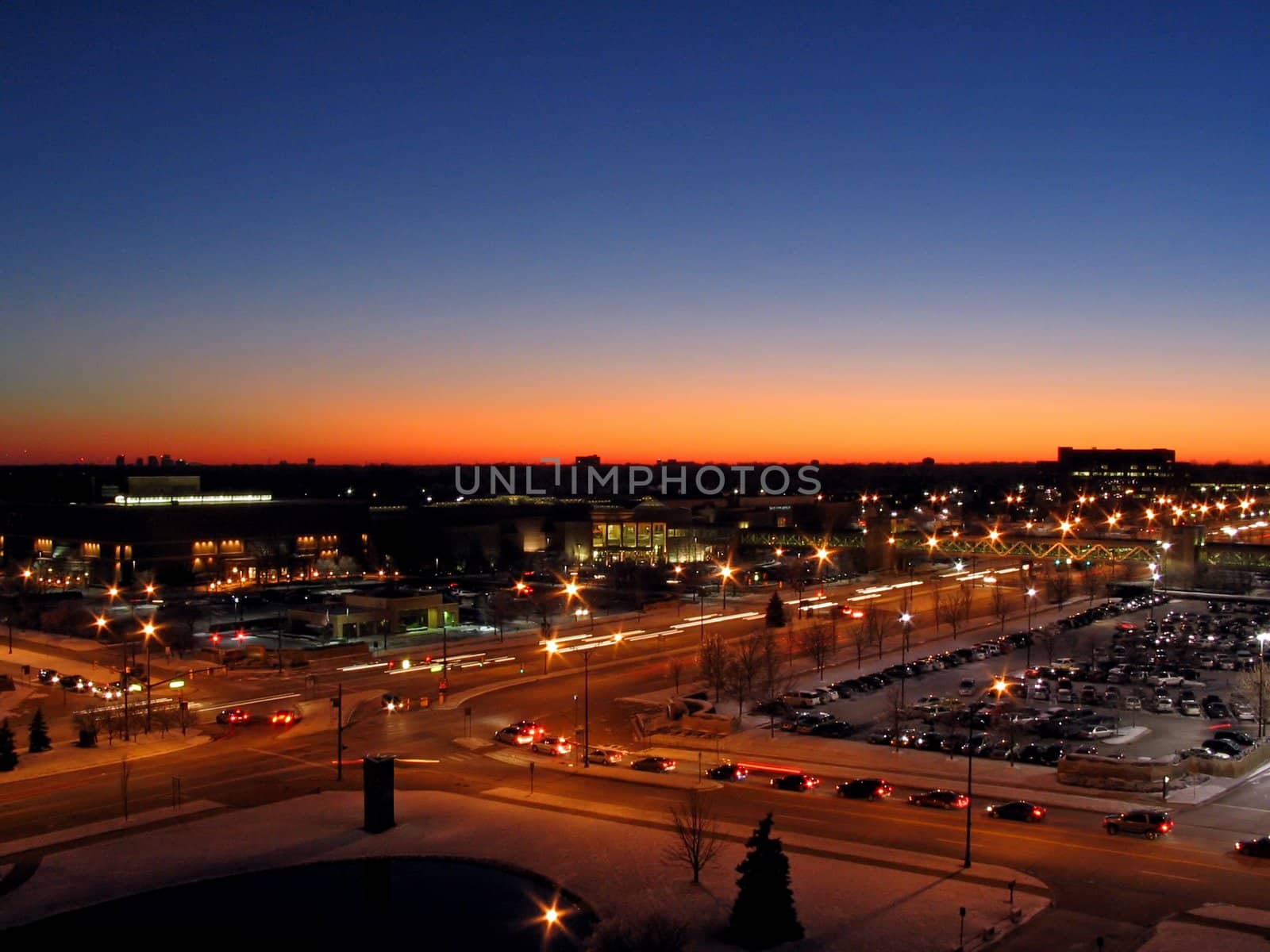 This is a night shot of a cross street in front of a shopping mall as the sun is setting in the background.