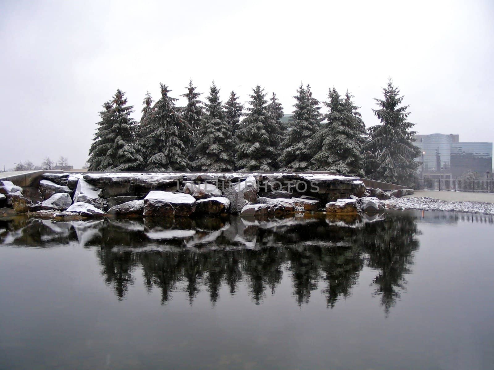 This is a picture of a small pond with pine trees and a office building in the background as snow is falling from the grey skies.