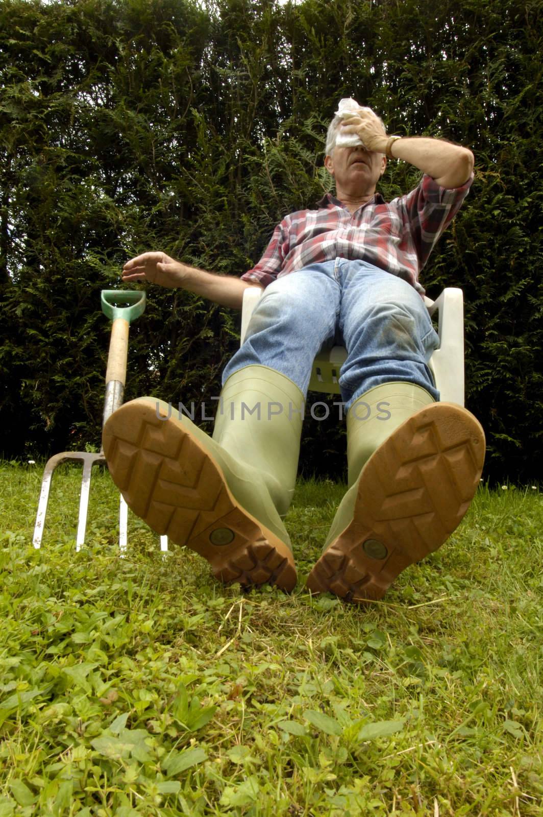 After a hard afternoon digging a gardener slumps back in a seat, exhausted, letting his fork fall to the ground, Space for text on the background to the left, or on the grass of the foreground.