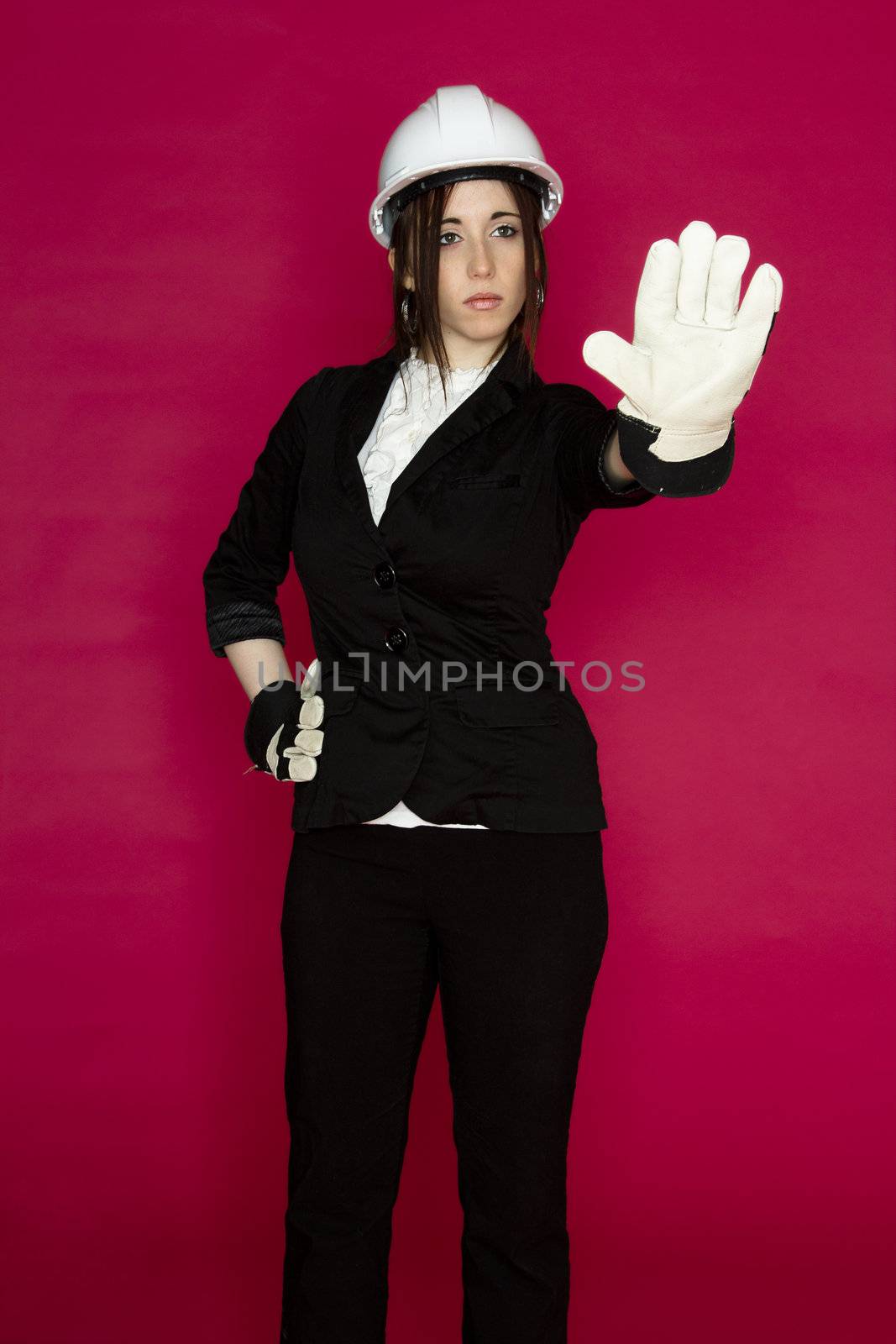 Young woman wearing white hard hat and construction glove, holding her hand up as a stop sign against a pink background