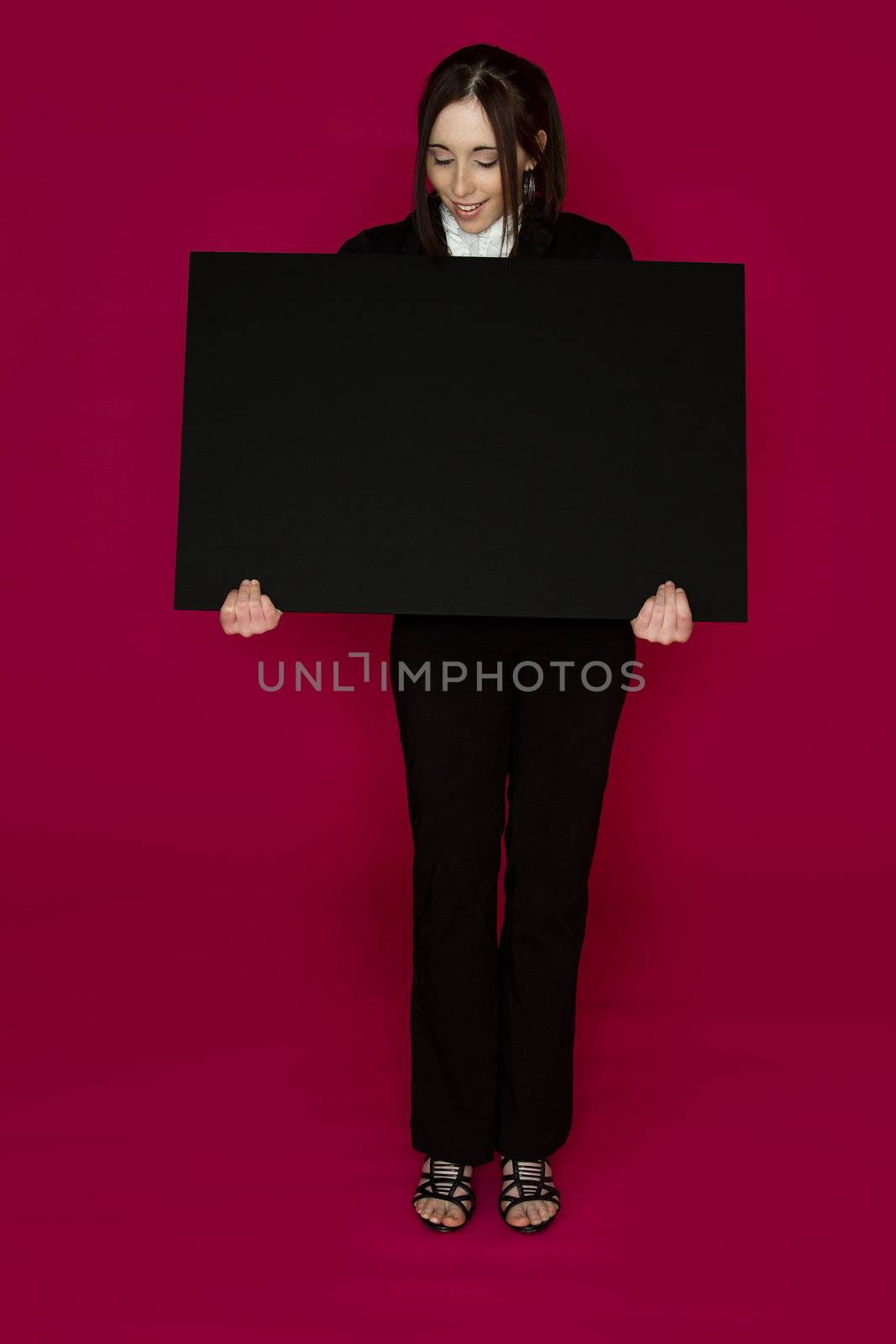 Young business woman wearing a business suit standing behind looking down at a black board against pink background