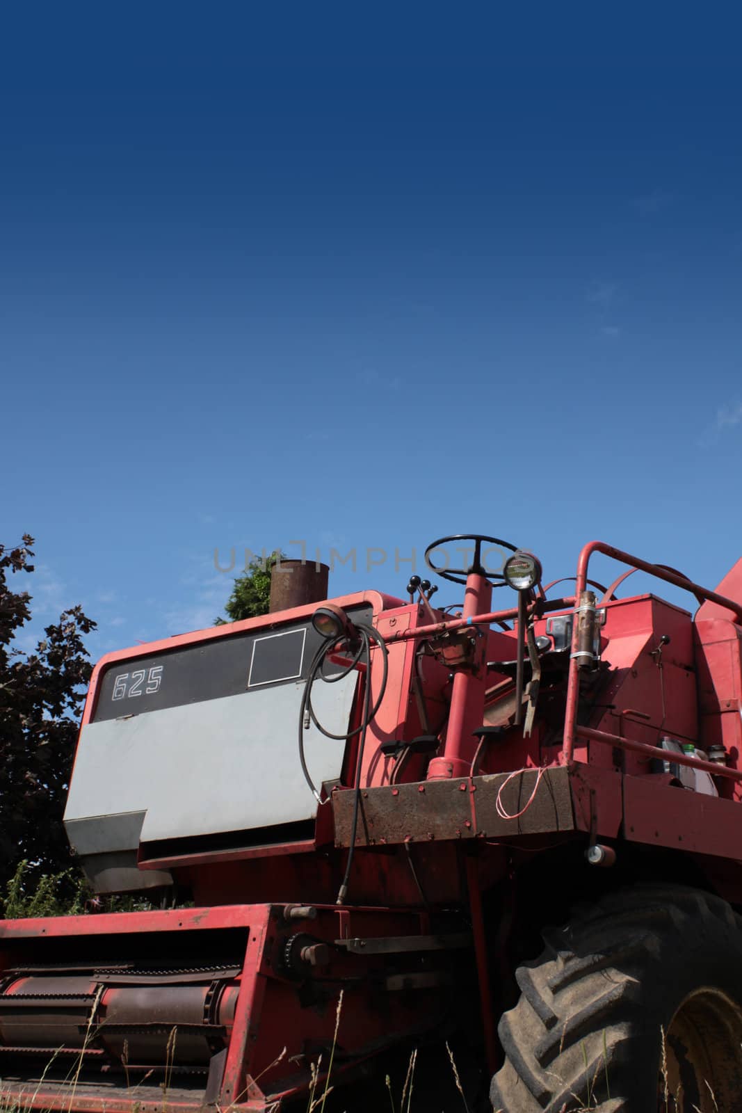 The front end of a red combine harvester machine stationary in a farm field in rural Wiltshire, England, set against a blue summer sky.
