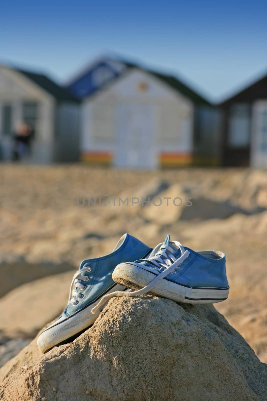 A pair of trainers/gym/beach shoes perched on top of some rocks at the beach, with beach huts in soft focus to the background.