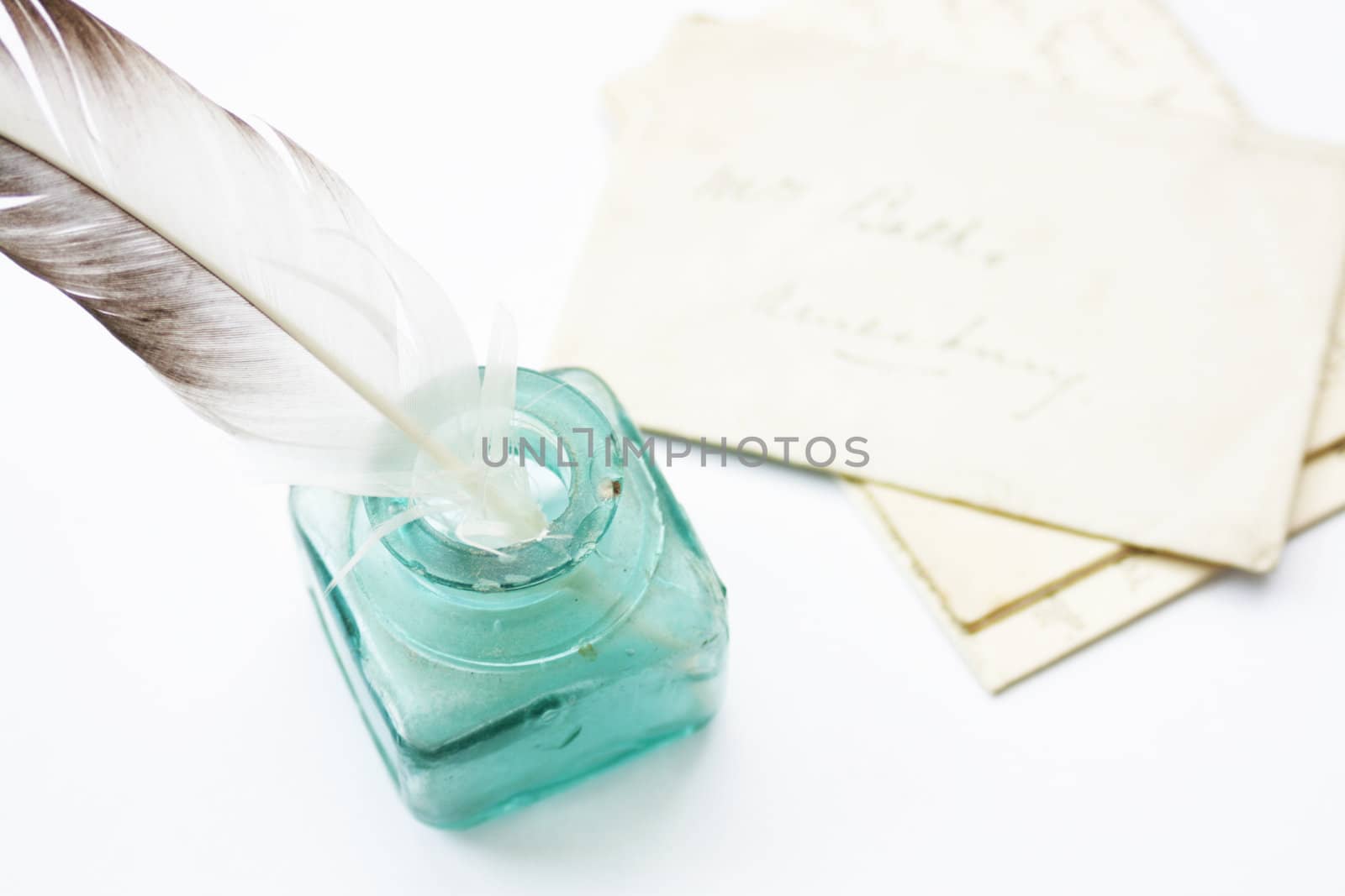 A feathered quill pen standing in a glass ink pot with old hand written letters behind, set on a white background