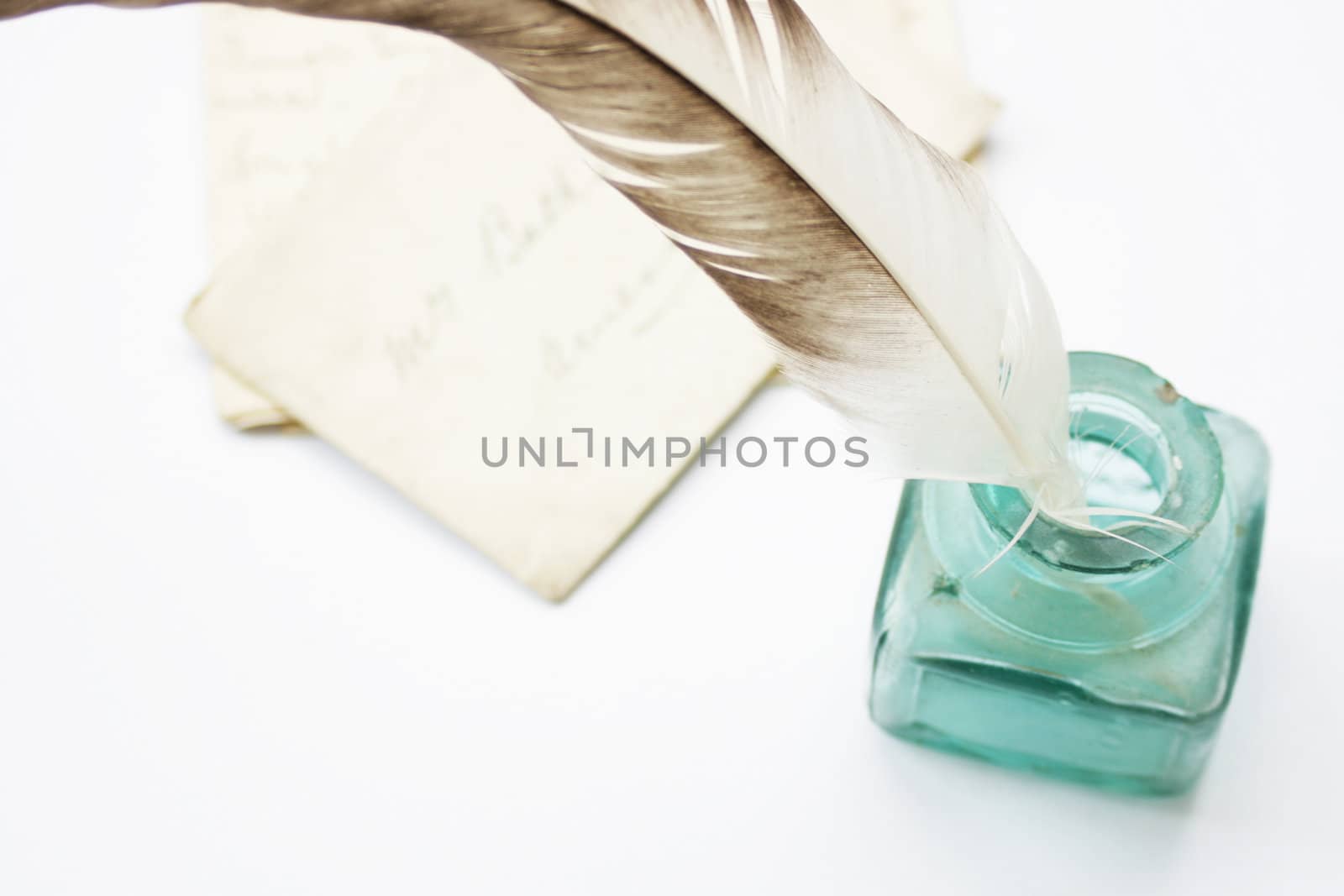 A feathered quill pen standing in a glass ink pot with old hand written letters behind, set on a white background