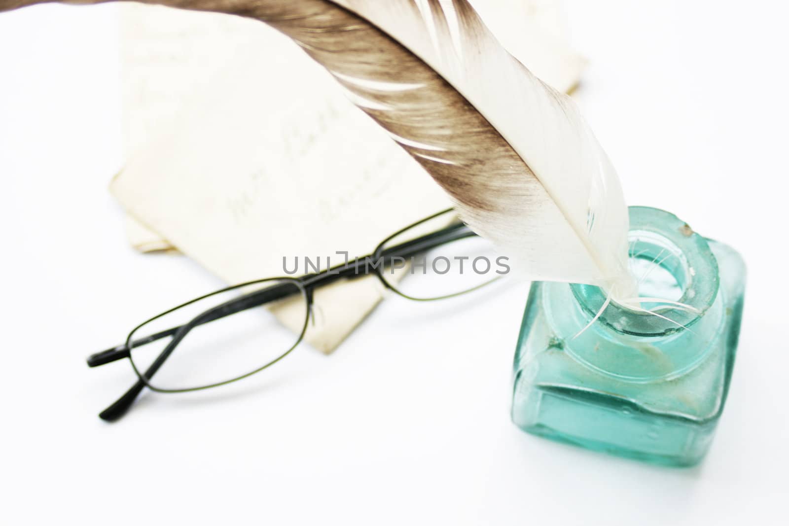 A feathered quill pen standing in a glass ink pot with old hand written letters behind with a pair of black framed reading glasses set on a white background