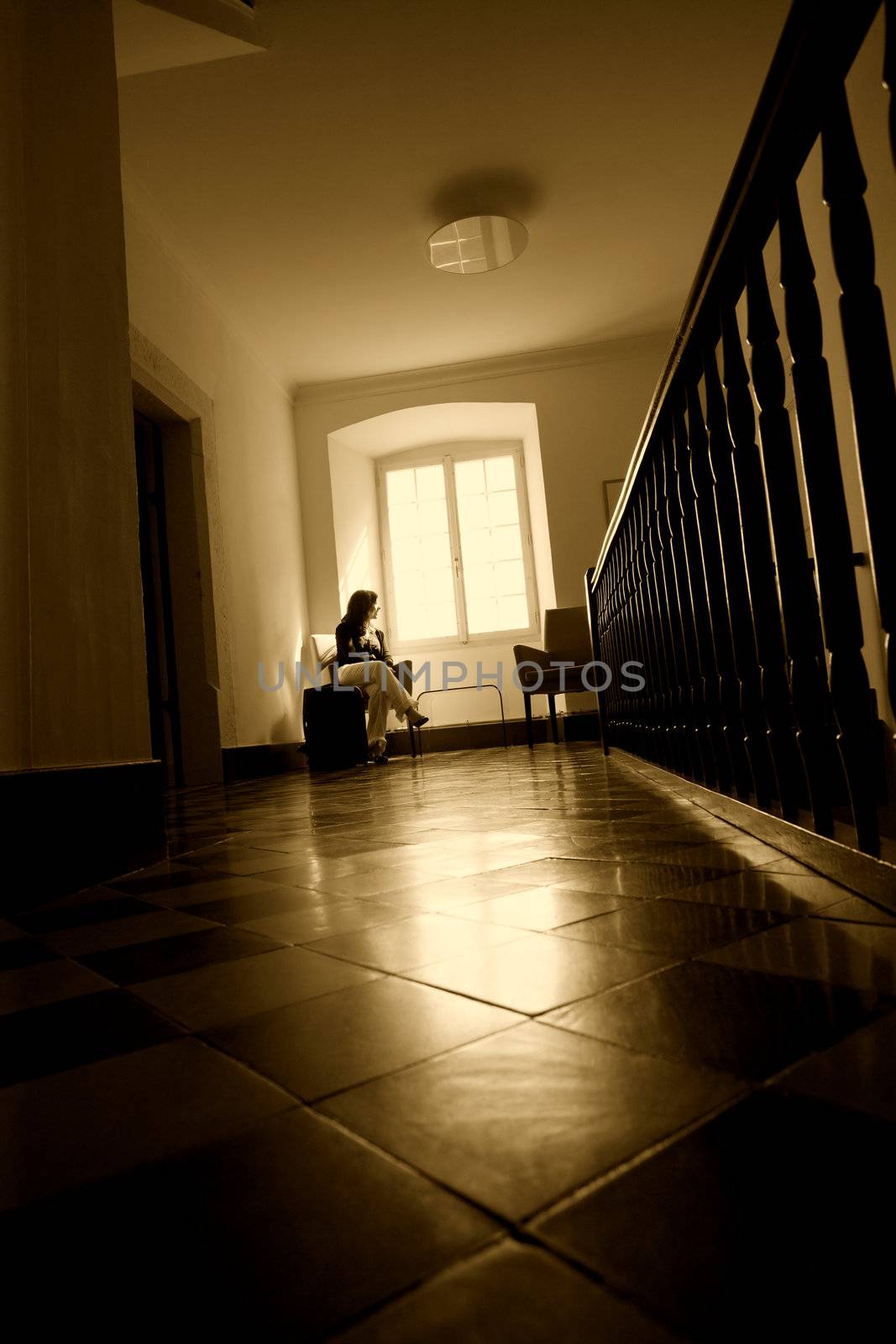 A beautiful young female waiting and looking out a large window. Suitcase by her side. Sunbeams coming through the window lighting the floor.