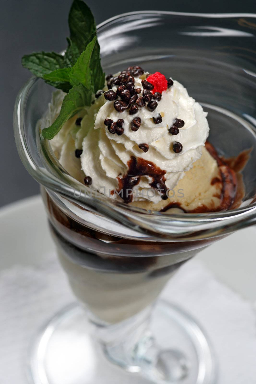 A closeup image of a vanilla ice cream sundae with whipped cream, chocolate covered nuts, chocolate sauce, and a raspberry on top.
