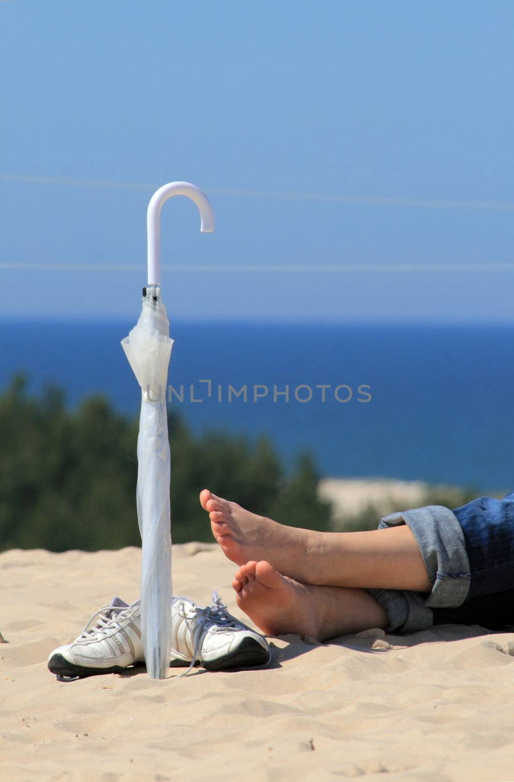 Woman's foot, pair of sneakers and umbrella on the sunny beach with the sea in background
