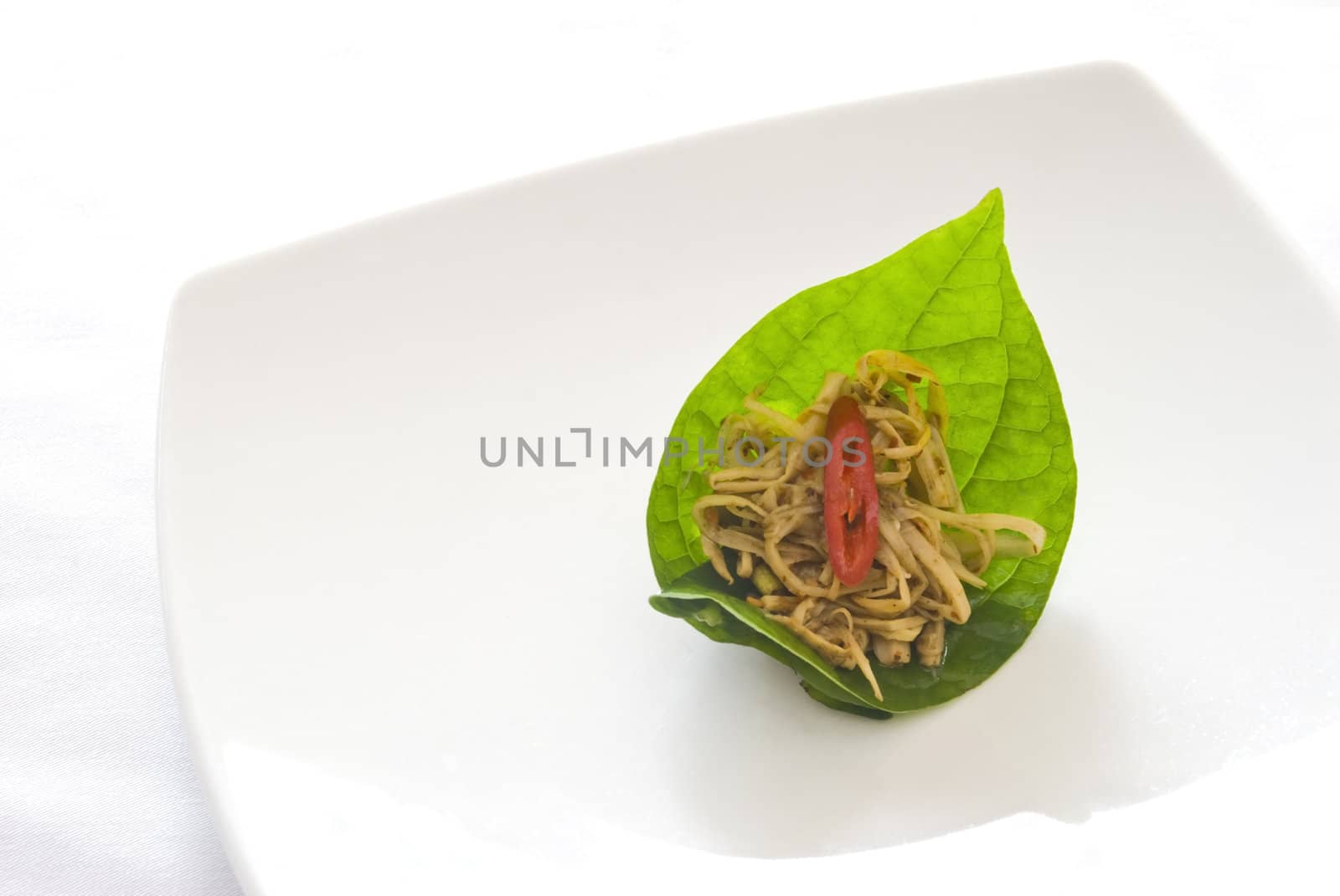 Spicy bamboo salad topped with a sliced red pepper, presented on a betel leaf and served on a white ceramic plate. Traditional Thai cuisine. 