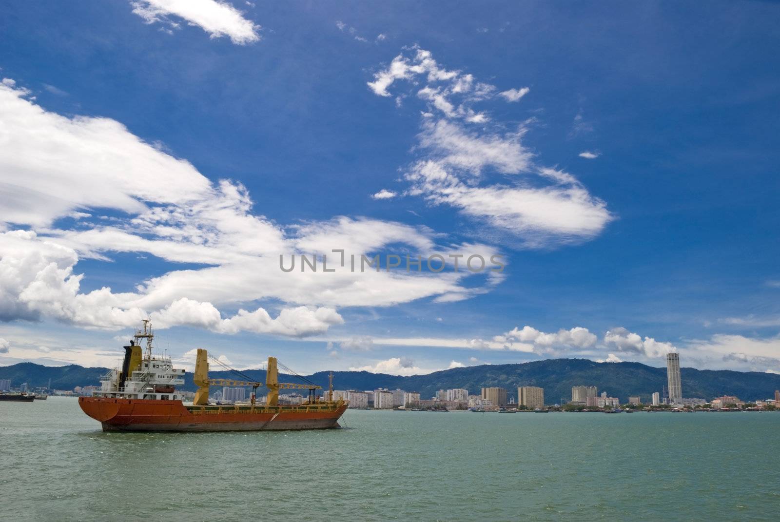 A large red cargo shipping vessel going through a strait and into the port of Penang in Malaysia. 