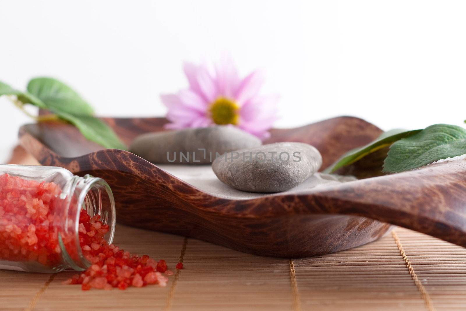 Massage stones on sand in a bowl, a purple flower and red bath salt isolated on white background