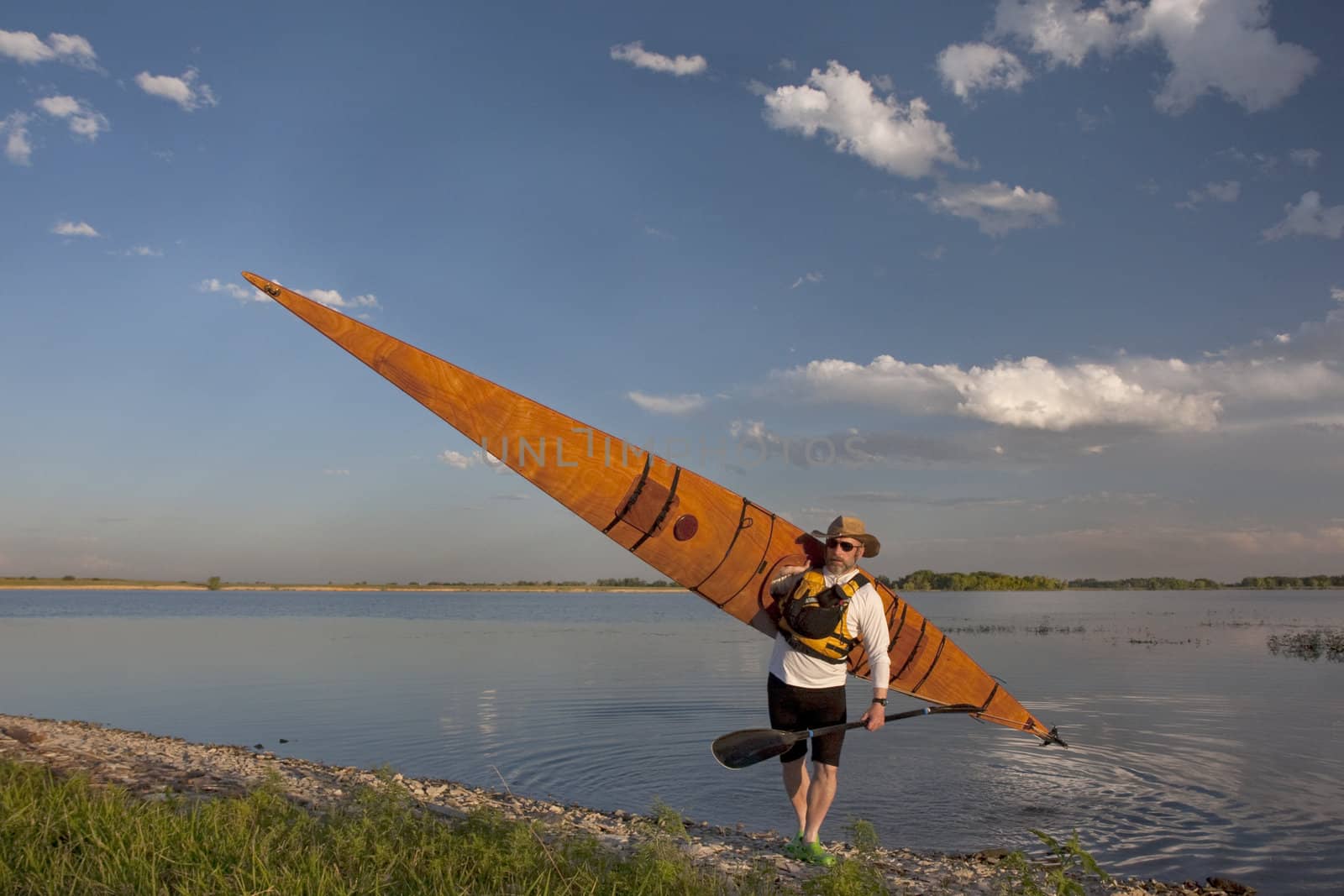 mature paddler in a hat and life jacket carrying his home built wooden sea kayak after paddling workout, grassy lake shore in northern Colorado