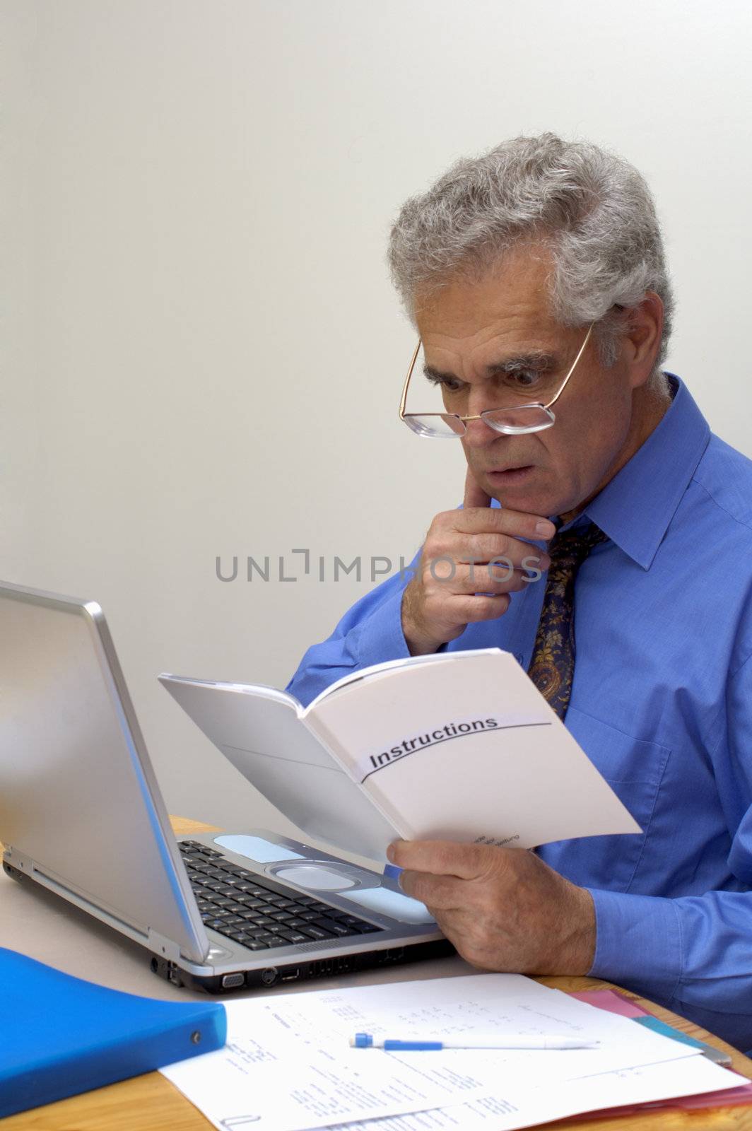An older businessman gazes in puzzlement at the instructions for his laptop computer, with a hint of worry on his face.