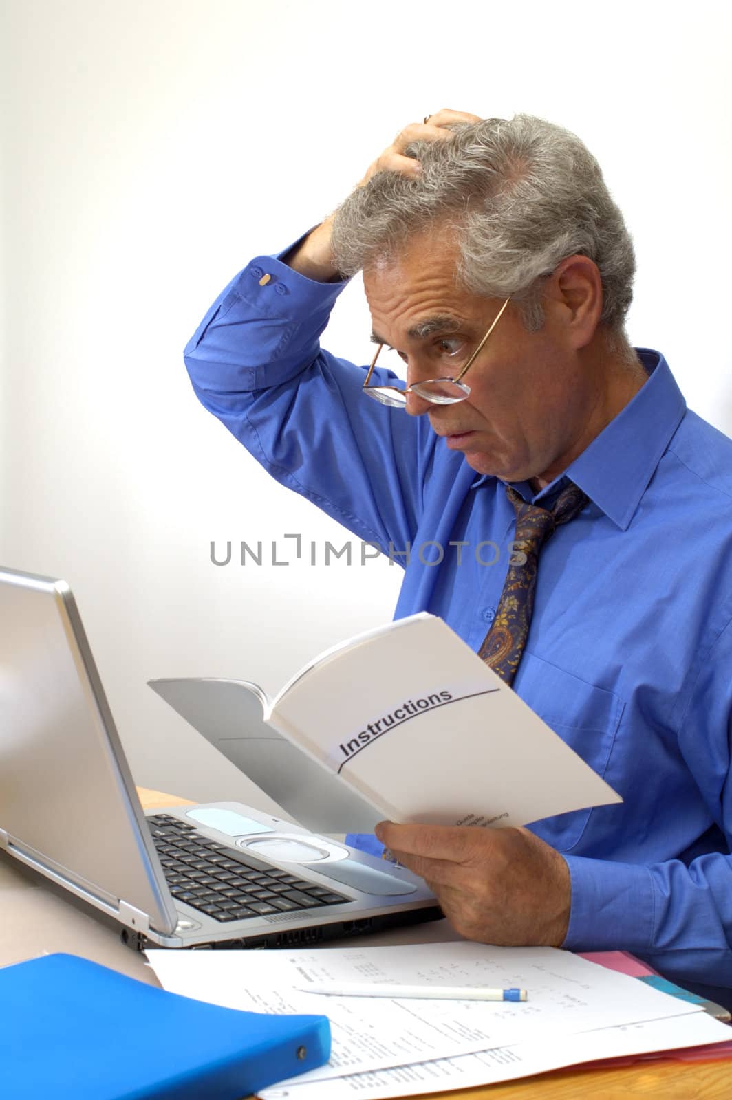 An older businessman gazes at the instruction booklet of his laptop, scratching his head in puzzlement. How does this thing work?