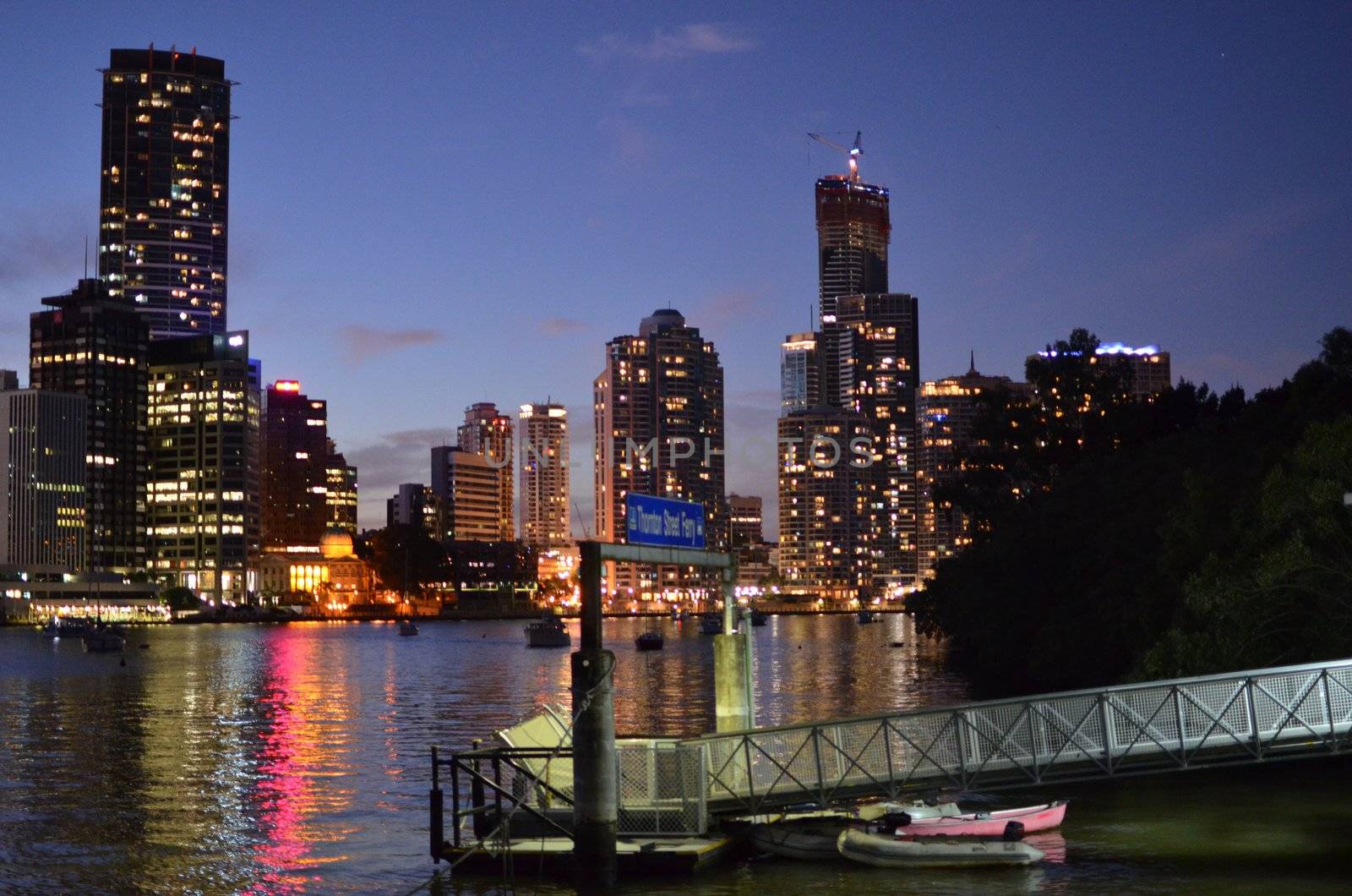 Cross river ferry jetty on the south bank of the Brisbane river in the foreground, with Brisbane city buildings and lights reflected in the Brisbane River behind.