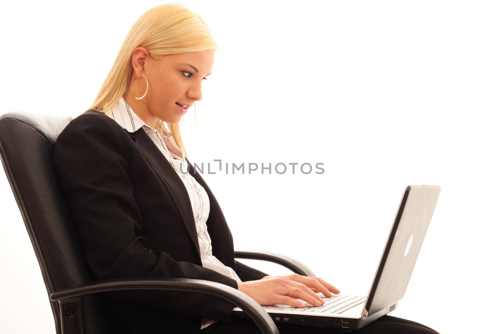 Young confident business woman with blonde hair, typing on a laptop in a stylish dark suit, sitting on a leather chair and a white background.