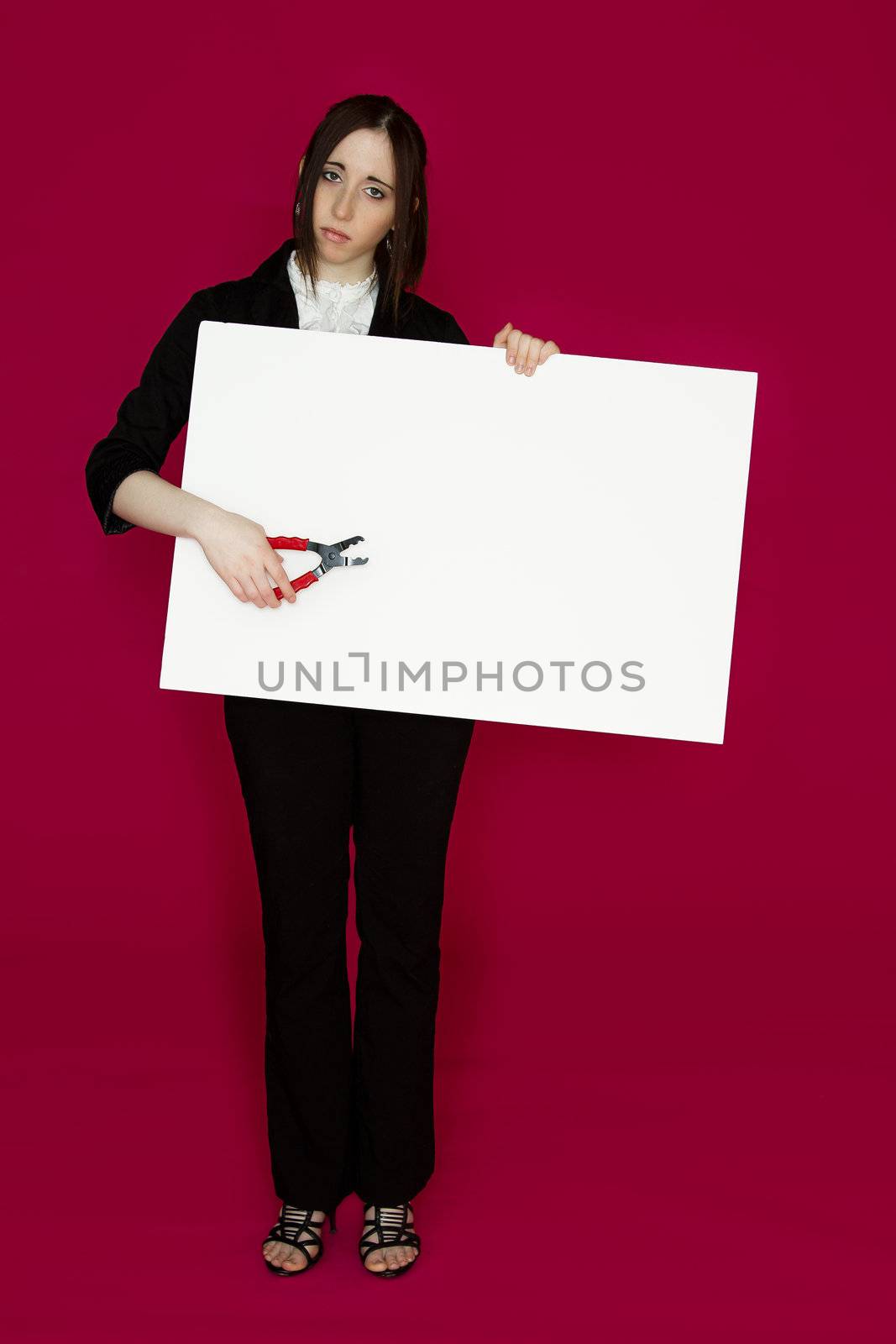 Young business woman holding a pair of pliers pointing to a white board with sad expression against pink background